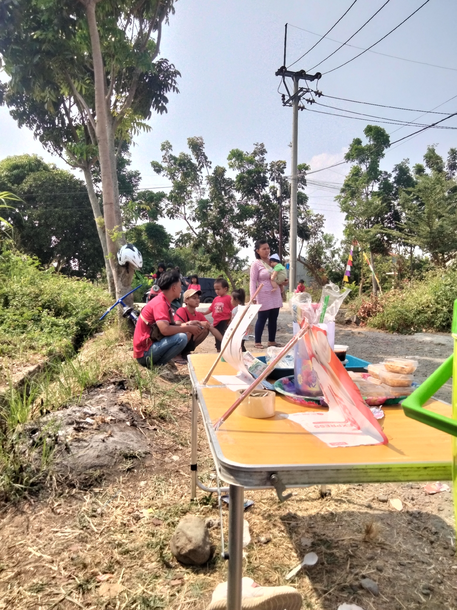 In Cariu, Indonesia, a group of people are gathered outdoors near a dirt road under a clear sky. In the foreground, a light-colored folding table is set up, laden with various items including food containers, plastic bags with utensils, and decorative flags. To the left of the table, a man in a red shirt and jeans squats, facing a group of children also wearing red shirts. A white and black helmet is attached to a tree trunk nearby.
Further back, a woman in a striped shirt holds a baby. Other children are scattered around, and a black vehicle is parked in the background. The environment is characterized by dry, grassy terrain, scattered rocks, and lush green vegetation. A utility pole with numerous wires stands prominently behind the gathering, indicating the presence of infrastructure. The overall scene suggests a casual outdoor event or picnic.