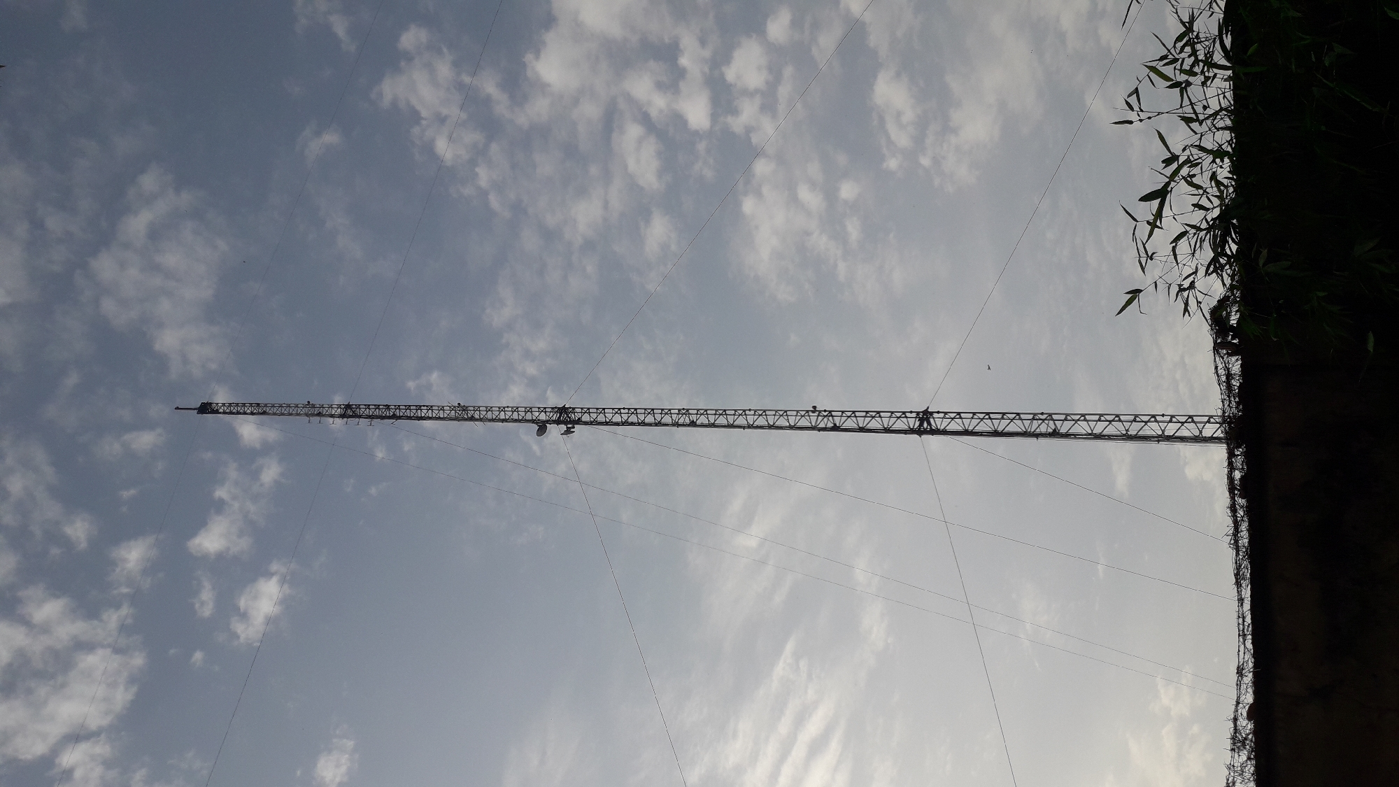 A tall metal tower stretches across a pale blue sky with scattered clouds. The tower is anchored by guy wires and has a lattice-like structure. Part of the right side of the image is obscured by dark foliage and a concrete or stone structure. The location is likely Onitsha, Nigeria, based on visual context and associated metadata. No people are clearly visible in the image.