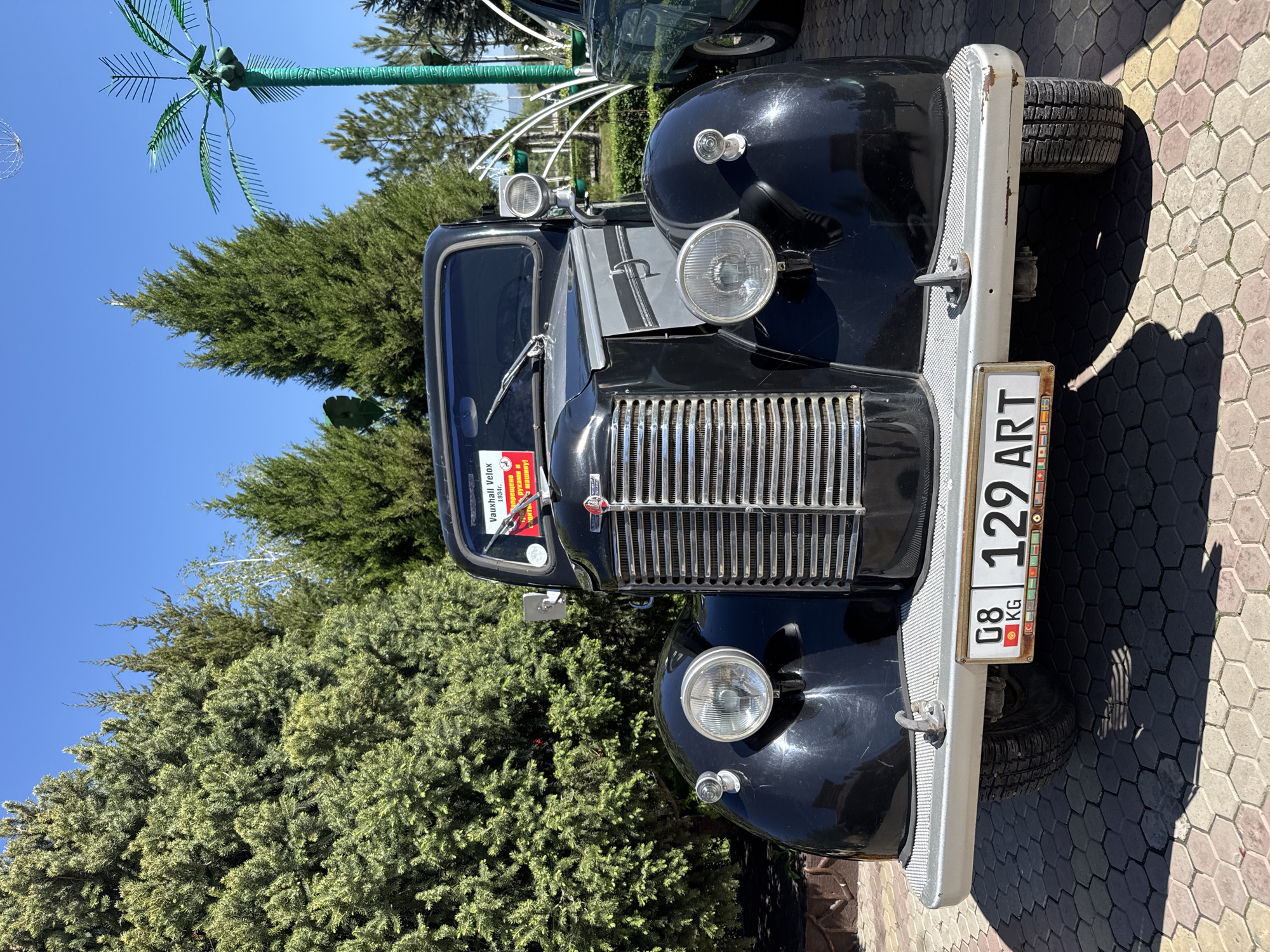 The image shows the front of a vintage black Vauxhall Velox car. The car features a chrome grill and bumper, as well as a license plate reading "08 KG 129 ART". It is parked on a paved surface with hexagonal patterns. Behind the car, there are green trees and an artificial metal palm tree. The sky is clear and blue. The scene appears to be in an outdoor setting, possibly a park or a display area, in the city of Kenbulun, Kyrgyzstan.