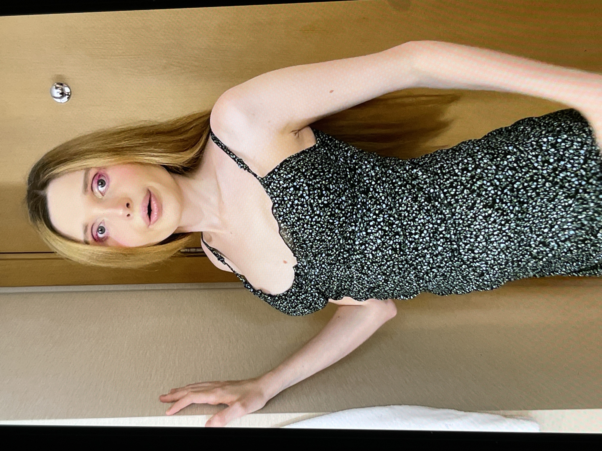 A full-body, medium shot shows a young, slender person with long, auburn hair, standing in what appears to be a hotel room. The person is wearing a dark, floral-patterned dress with thin straps and a scalloped neckline. Their face is turned slightly towards the camera, with striking pink eyeshadow applied to their eyelids. Their mouth is open, and their eyes are wide. The person is leaning their left arm against a textured, beige wall on the left side of the frame. To the right of the person is a closed wooden door with a silver knob. The background is simple and uncluttered, suggesting an indoor setting.