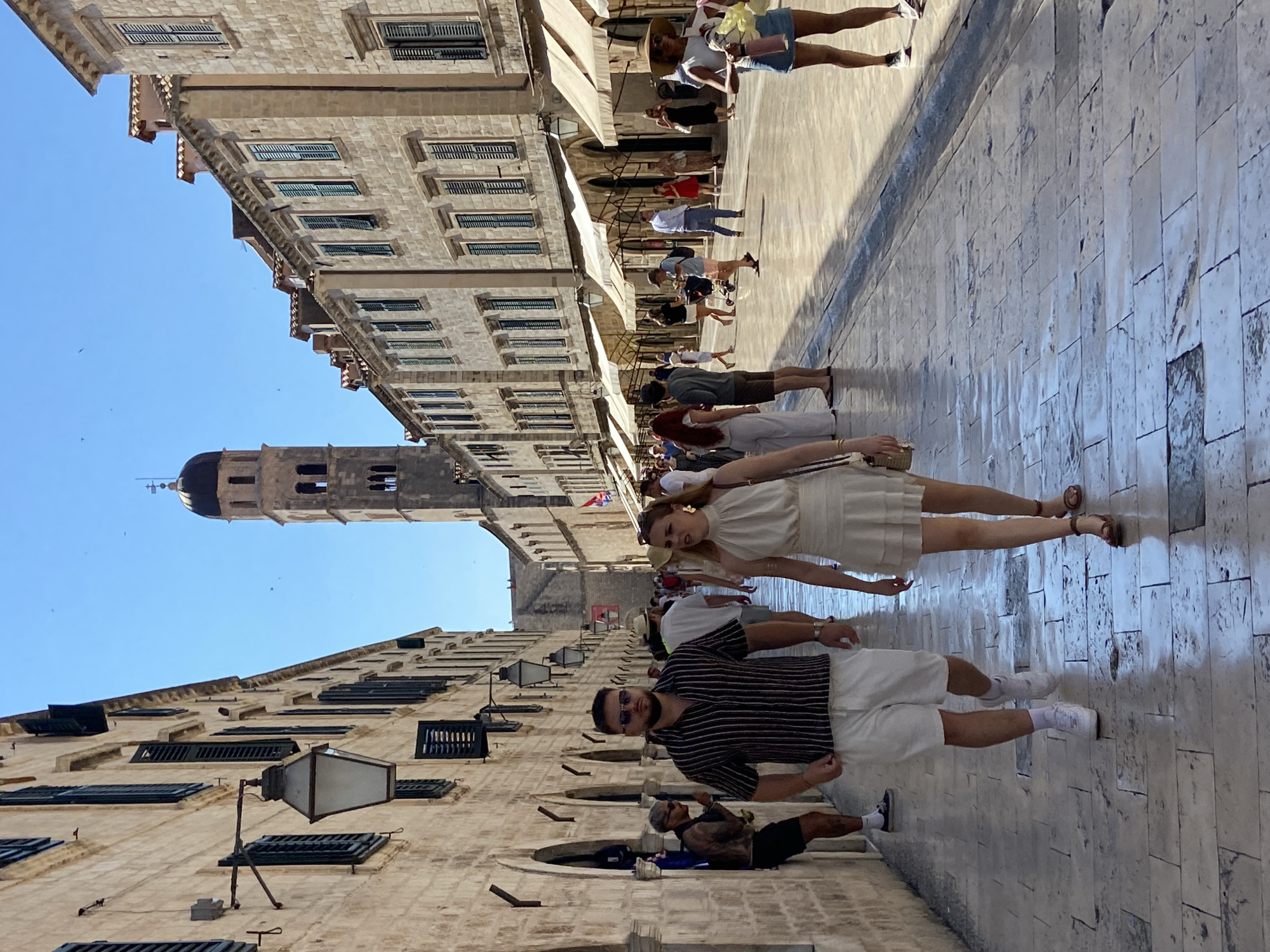 The image depicts a busy, sunlit street in Dubrovnik, Croatia, likely the Stradun, the main street of the Old Town. A young couple walks towards the viewer in the foreground. The man wears a black and white striped short-sleeved shirt and white shorts, with white sneakers and sunglasses. The woman wears a cream-colored sleeveless dress with ruffles and brown sandals. Behind them, a diverse crowd of tourists and locals stroll along the wide, paved street. The architecture is characteristic of Dubrovnik, with stone buildings lining both sides of the street, featuring arched windows, shuttered windows, and awnings above shop fronts. In the distance, a tall bell tower with a dark dome stands against a clear blue sky. The stone pavement is wet in places, suggesting recent rain or cleaning. The overall scene is one of vibrant daytime activity in a historic European city.