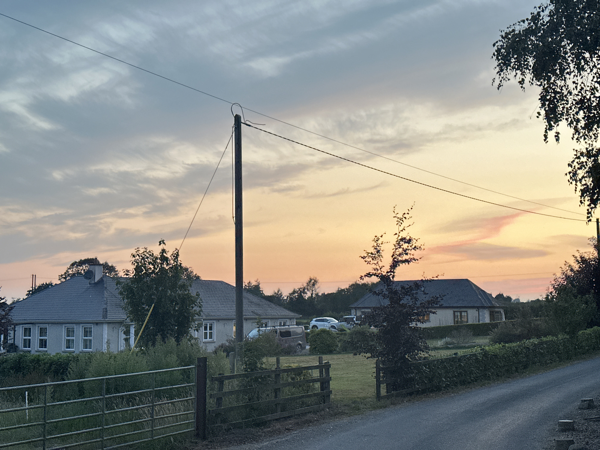 The image depicts a rural landscape at dusk in Moortown, Ireland. A grey asphalt road curves into the foreground, bordered by a low wooden fence and dense green foliage. Beyond the fence, a spacious, detached house with a grey tiled roof is visible, featuring multiple windows. In the middle ground, another similar house with a grey roof is situated, with several vehicles parked in its driveway, including a white SUV and a dark minivan. Trees and a utility pole with wires are scattered throughout the scene. The sky is a gradient of soft blues, oranges, and pinks, indicating the setting sun.