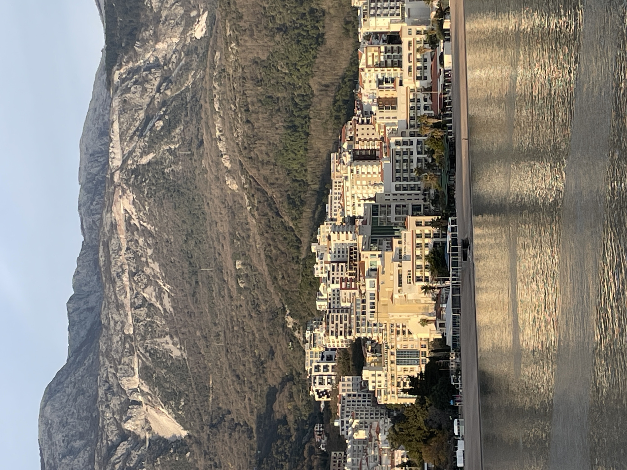 The image depicts a panoramic view of the coastal city of Budva, Montenegro, specifically focusing on its built-up area and the adjacent mountains, observed across a body of water.
In the foreground, a calm expanse of water, likely the Adriatic Sea, occupies the lower third of the frame. Sunlight reflects off its surface, creating shimmering, elongated golden patterns.
Immediately beyond the water, a sandy beach is visible, running horizontally across the mid-ground. On this beach, a single, small, dark human figure stands near the water's edge, facing the city. Several low-rise structures are positioned on or directly behind the beach: notably, a series of white, tent-like shelters to the left and a longer, enclosed building with a dark roof and large windows, possibly a restaurant or beach club, extending towards the right. A small, dark, rectangular object, possibly a raft or small boat, is partially submerged near the shore.
Behind the beach and its immediate structures, the urban landscape of Budva rises. A dense array of multi-story buildings, predominantly white, beige, and light-colored residential or hotel structures, cascade up the lower slopes of the background mountains. Many buildings feature balconies and varied architectural details; some display visible signage, including "HOTEL SPLENDID" partially discernible on one prominent structure. Green foliage, including palm trees, is interspersed among these buildings. The structures are illuminated by warm, golden light, indicative of either sunrise or sunset, creating elongated shadows.
The background is dominated by a large, steep mountain range. The lower sections are covered in dense, dark green deciduous and coniferous trees. Higher elevations reveal extensive grey and white exposed rock faces, marked by geological features, evidence of erosion, and visible human alteration such as quarrying or road construction scars. The sky above the mountains is a clear, pale blue.