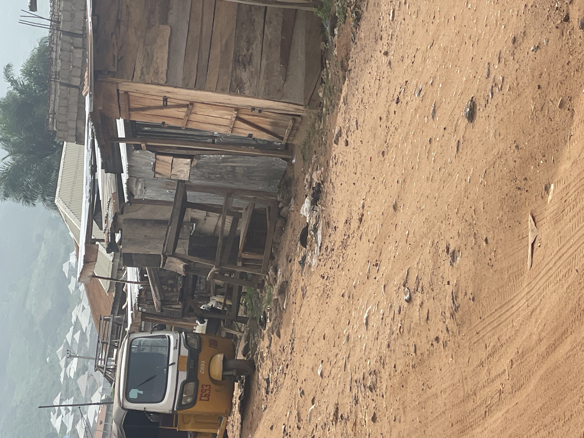 This image depicts a dusty street scene in Lokoja, Nigeria, with a prominent yellow auto-rickshaw, locally known as a "Keke Napep," parked on the left. The vehicle displays the registration number "C693." In the background, various makeshift structures constructed from corrugated metal, wood, and concrete bricks line the street, suggesting a commercial or residential area. The environment is characterized by arid, reddish-brown earth, with scattered rocks and debris, indicative of a dry season. The sky appears overcast, with a hazy, muted light. In the distance, a cluster of buildings with white roofs and tiled structures can be seen ascending a hill. The overall impression is one of a bustling, yet modest, urban landscape in Nigeria.