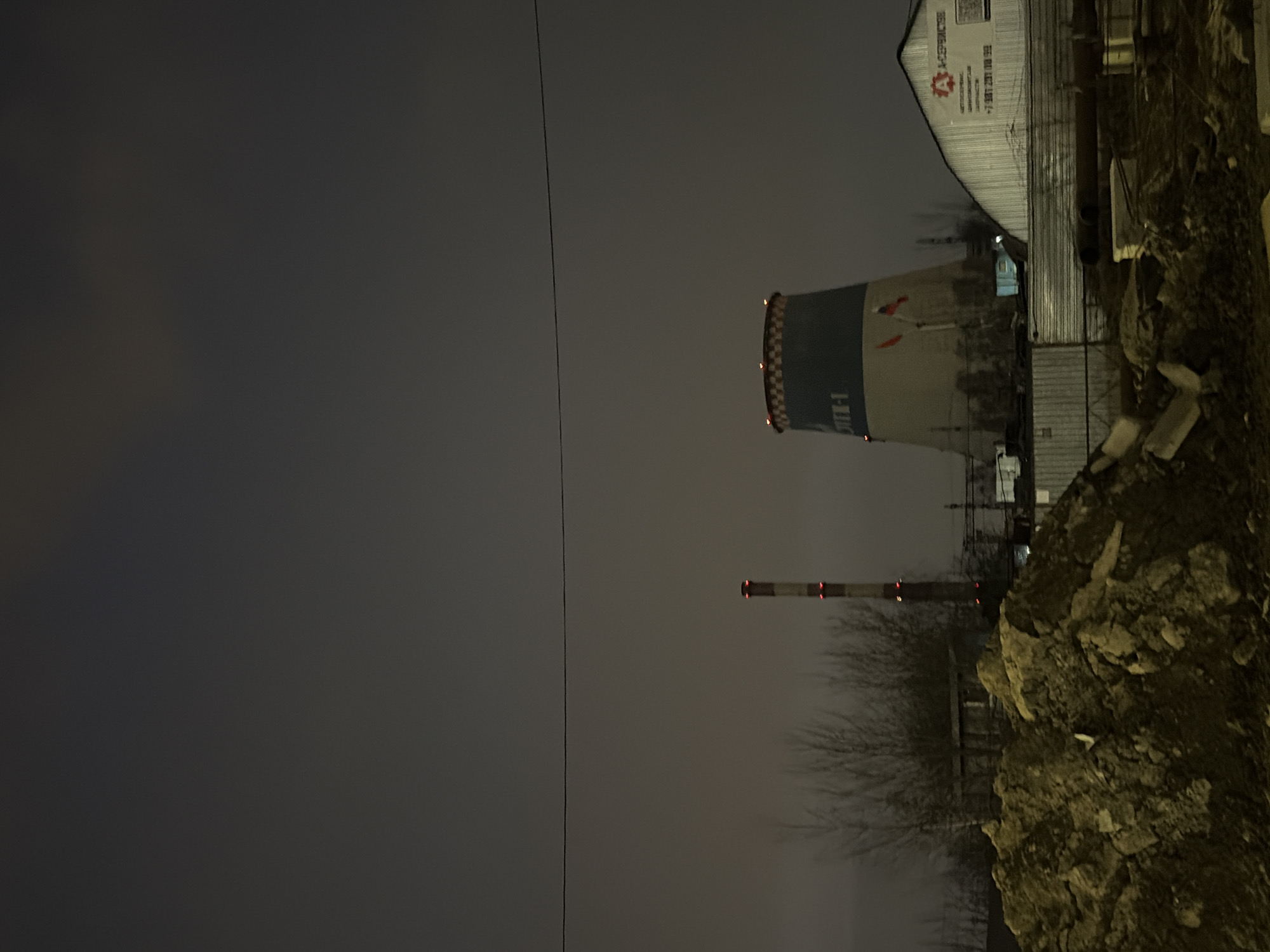 The image depicts an industrial scene at night under a dark, overcast sky. The location is TETS-1 (Thermal Power Plant No. 1) in Saint Petersburg, Russia.
In the mid-ground, a tall, slender chimney, striped reddish-brown and light gray, is visible to the left of the center, featuring two bands of red aviation warning lights on its upper half. To its right stands a large, hyperbolic cooling tower, predominantly light gray or off-white at its base, transitioning to a dark blue or dark gray section with the Cyrillic text "ТЭЦ-1" clearly visible. The top rim of the cooling tower is adorned with a checkered pattern of light and dark squares, likely lights. Two small Russian flags are mounted on poles extending from the lower-mid section of the cooling tower.
Bare, deciduous trees are sparsely scattered between the foreground and the industrial structures. A horizontal corrugated metal fence, topped with barbed wire, separates the foreground area from the power plant facilities. Behind the fence and to the far right, a light-colored, single-story building with a sloped roof displays a red logo with a stylized 'A' and the Cyrillic text "А-СЕРВИССТРОЙ" (A-ServiceStroy), along with a QR code and a Russian phone number: "+7 981 731 00 32". Dark, cylindrical objects, resembling pipes, are visible on the ground behind the fence.
The foreground is dominated by large, uneven piles of excavated earth and rocks, appearing dark brown and yellow-brown due to the limited ambient light. Various pieces of concrete debris and other construction materials are scattered amongst these piles. A single, dark overhead wire extends horizontally across the upper-mid portion of the frame. No individuals are visible in the scene, and no overt actions or interactions are depicted. The scene is illuminated by artificial light sources originating from the industrial structures, casting a dim glow on the immediate surroundings.