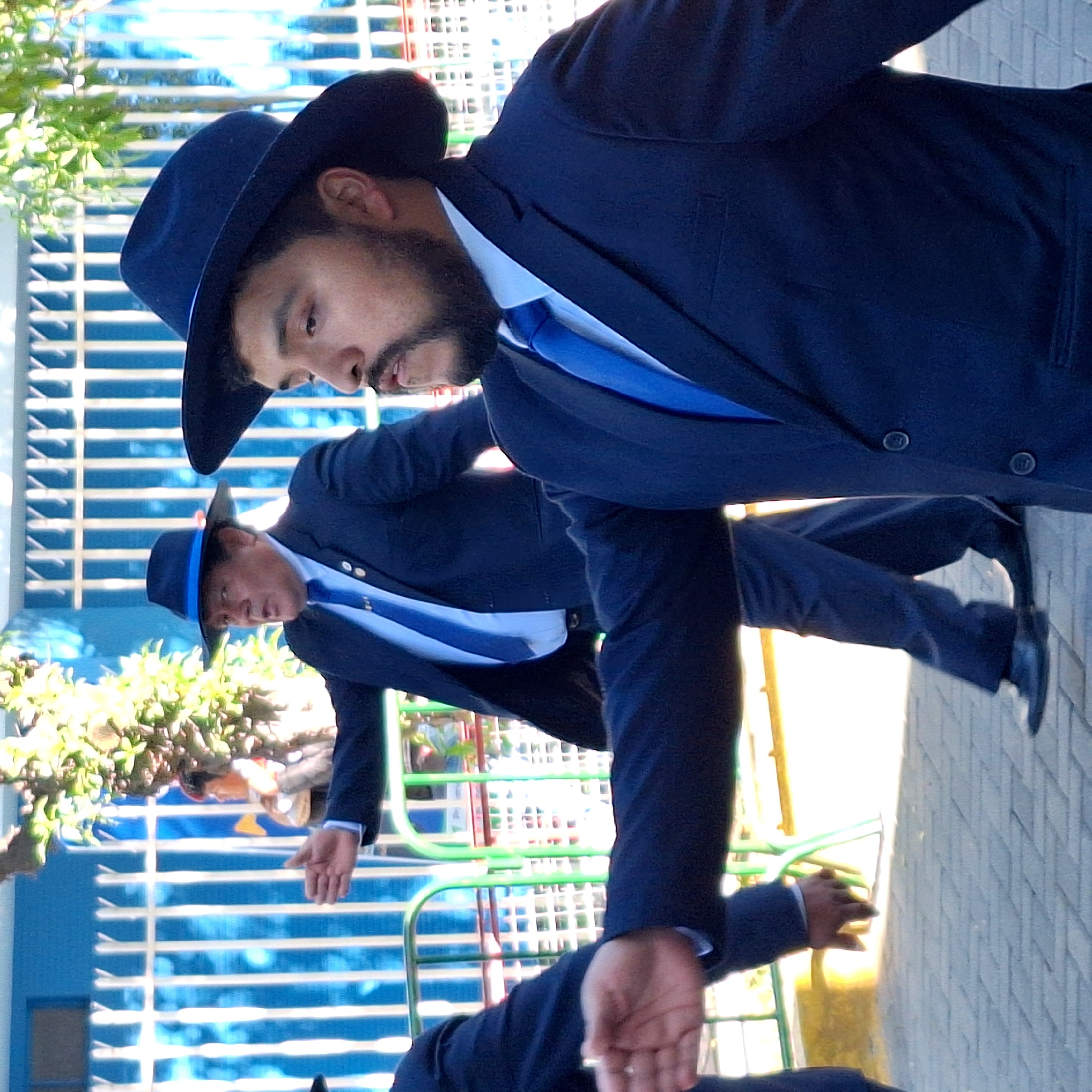 Two men in Cerro Colorado, Peru, wearing matching dark blue suits, light shirts, and blue ties are performing a synchronized dance move, extending their hands. Both men are wearing wide-brimmed dark hats. The background includes a blue and white fence, greenery, and a paved walkway. A third man in a suit is partially visible in the foreground.