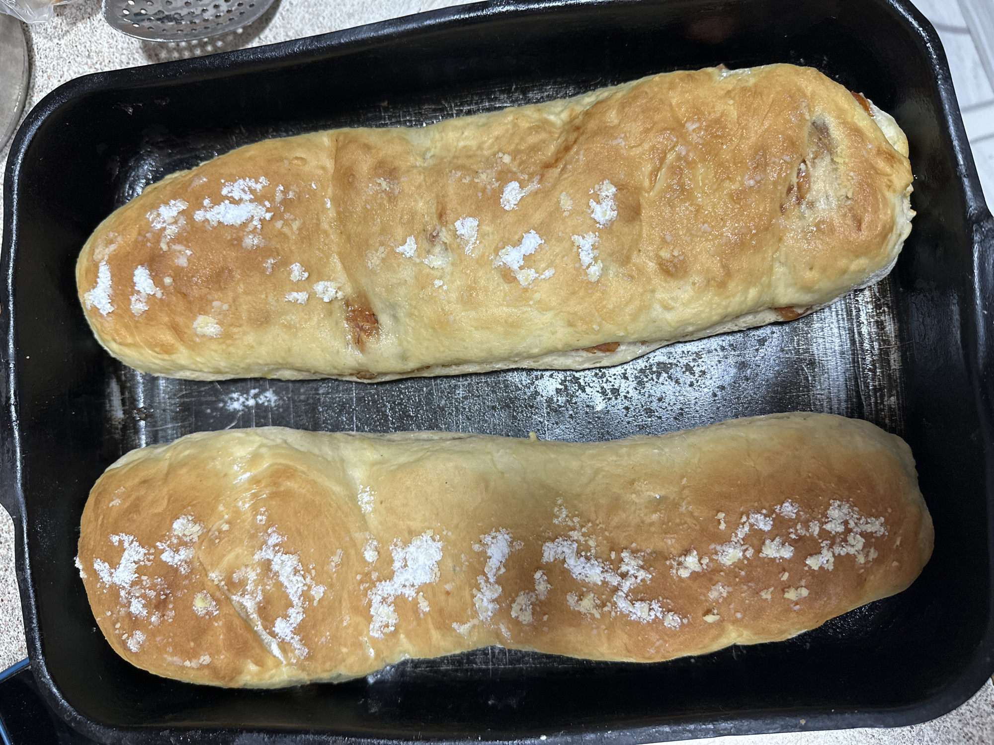 Two loaves of baked bread are in a black baking pan. White powder is sprinkled on top of the bread. A slotted spoon is visible in the upper left corner. The location is not identifiable from the image.