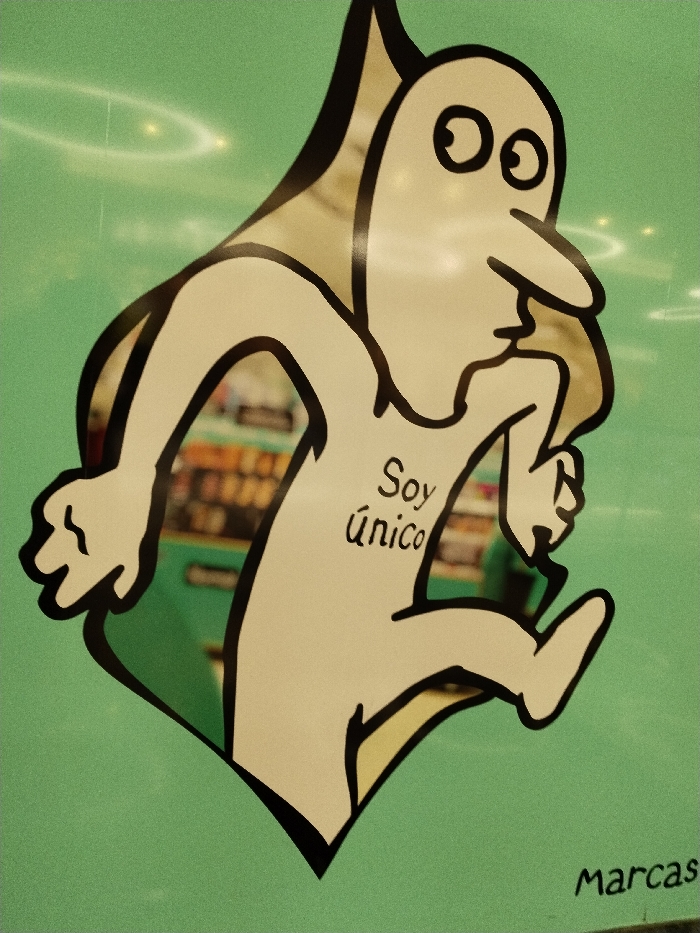 A cartoon-like figure on a green background is running. The figure is primarily white with a black outline and has a large nose, round eyes, and a head that extends upwards. On its torso, the Spanish phrase "Soy único" is written in black script. The running figure is framed by a black, leaf-like shape. The word "Marcas" is written in the lower right corner of the image. The background is a solid, pale green color with faint, light-colored, ring-like shapes. The figure appears to be a sticker or poster adhered to a reflective surface, capturing a blurred interior scene within its borders.