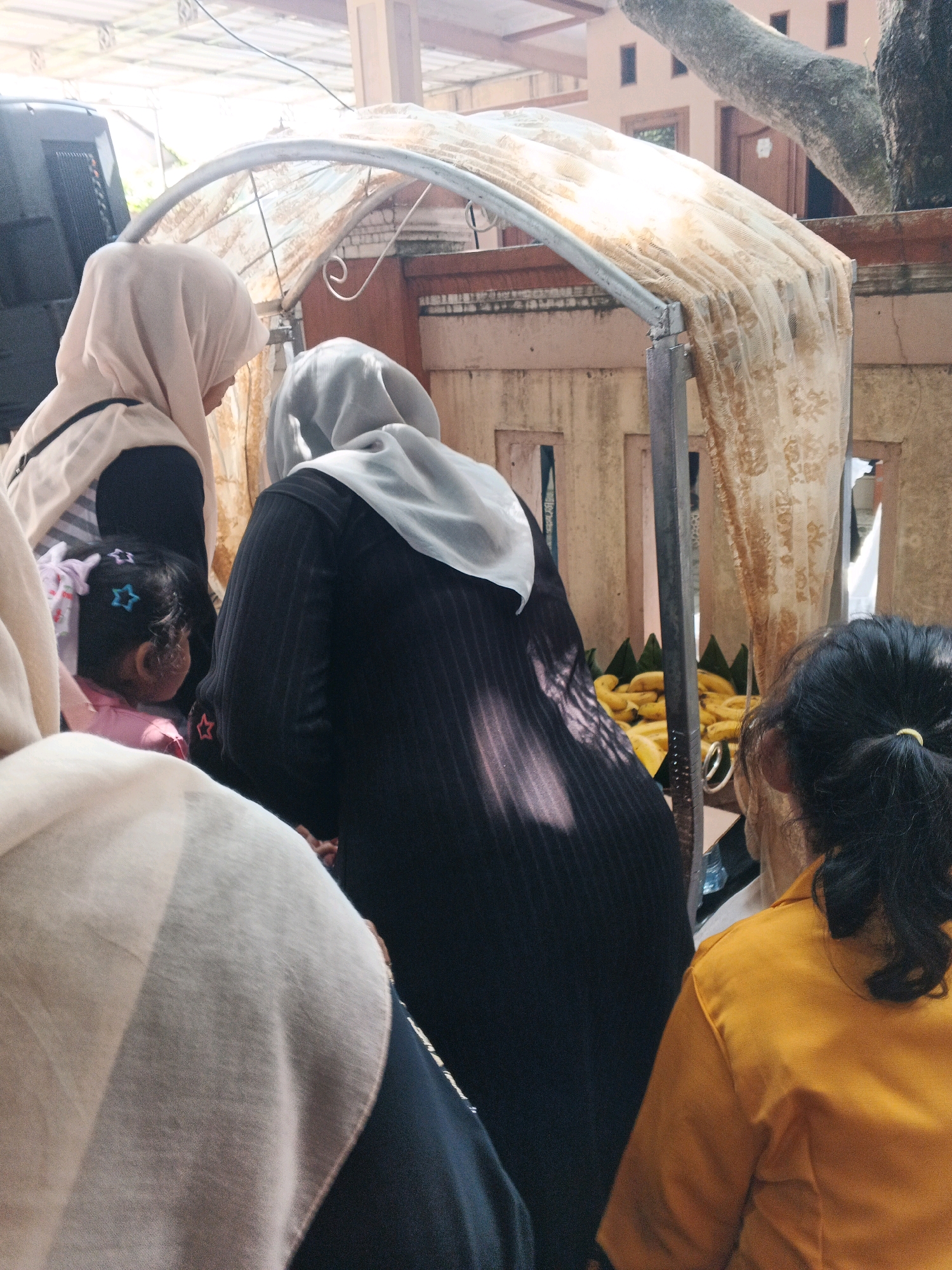 This image depicts an outdoor scene, possibly a market or gathering, in Kadu, Indonesia. In the foreground, several individuals are partially visible, wearing headscarves and traditional clothing. One individual is wearing a light-colored headscarf and a dark garment, while another is in a black striped dress with a light-colored headscarf. A young child with star-shaped hair clips is seen in the center. Another person in the foreground is wearing a bright yellow garment.
Behind the people, there is a stall or structure covered with a sheer, patterned fabric, illuminated by sunlight. A large tree trunk is visible to the right, with part of a building in the background. A pile of bananas is displayed on the stall, suggesting it might be a food vendor. The overall atmosphere appears to be casual and communal, with people interacting and possibly shopping. The setting is characterized by natural light and the presence of both human activity and natural elements.