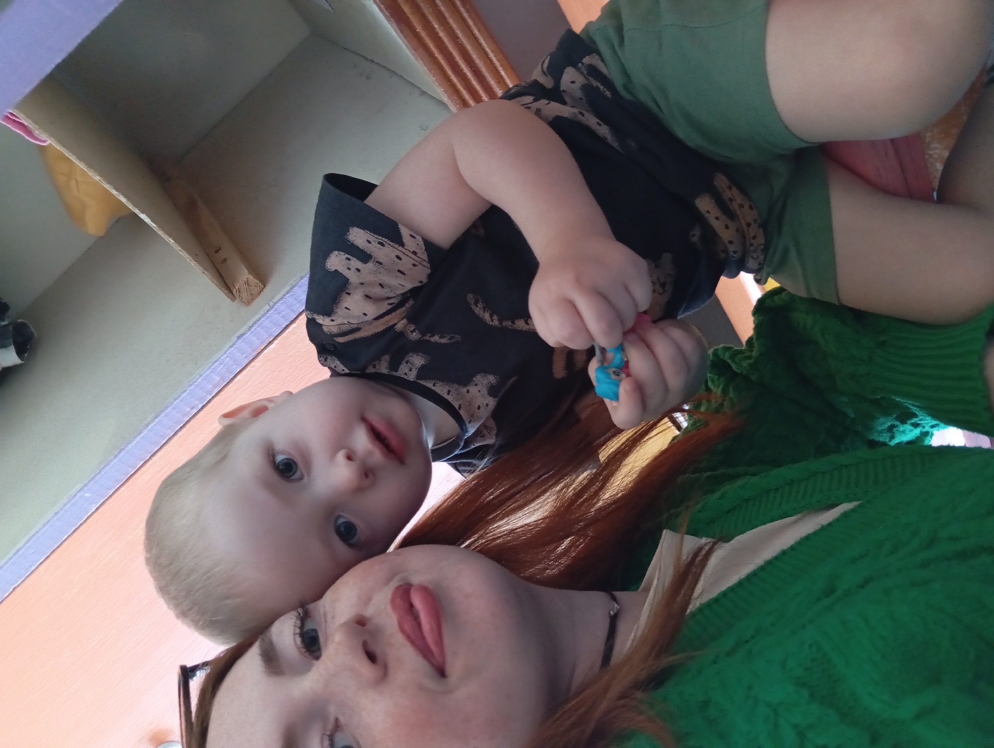 A selfie shows a woman and a child. The woman has red hair and freckles. She is wearing glasses and a green sweater. The child has blonde hair and blue eyes and is wearing a dark shirt with animal prints and green shorts. The child is holding a toy in his right hand. The background shows a shelf and a curtain. It is impossible to confirm the specific location in the city of Yurgamysh, Russia from the available image.