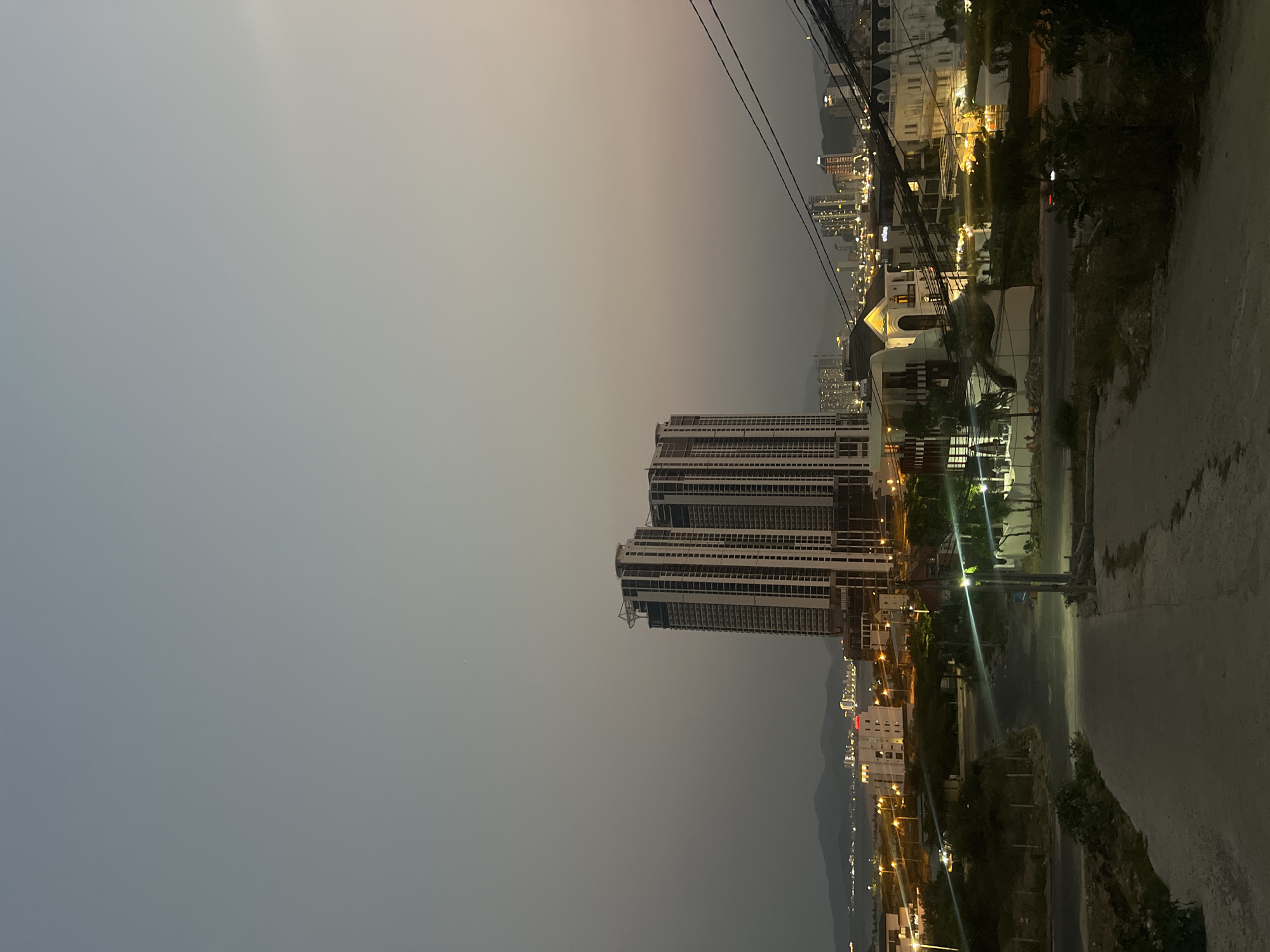 The image shows a cityscape in Bắc Nha Trang, Vietnam, taken at twilight. A tall condominium building, under construction, is the central focus. To the right, there is a roadway, vegetation, and other residential structures with lit windows. Utility lines are visible near the top of the frame. Lights of other buildings are visible across the water, where mountains are also. The sky is gray, with a gradient towards lighter colors at the horizon.
