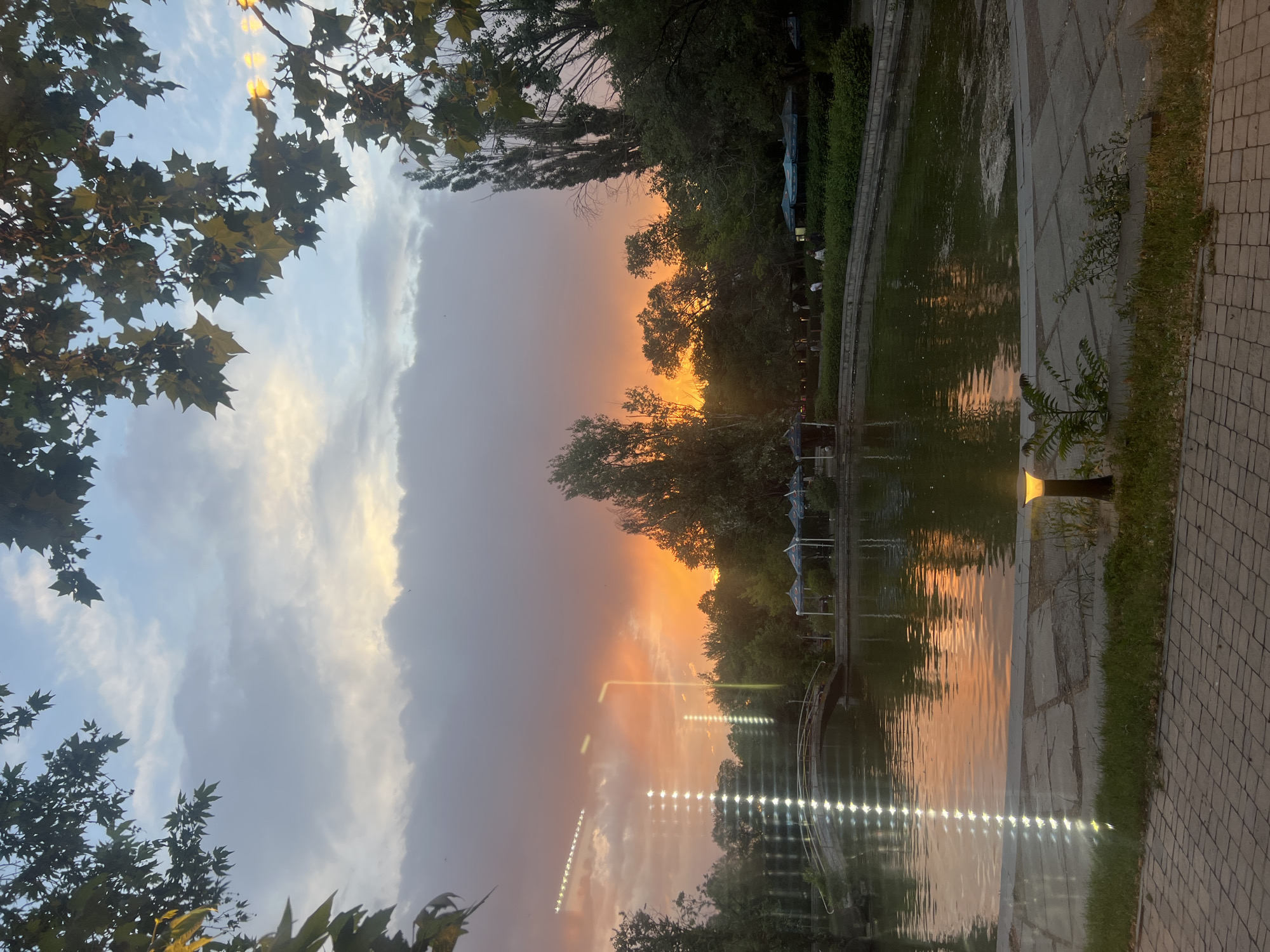 The image depicts a serene park scene in Yerevan, Armenia, during sunset. A tranquil lake occupies the foreground, reflecting the vibrant orange and pink hues of the sky. A charming bridge spans a section of the lake, adding to the picturesque setting. Lush green trees and foliage surround the lake, with some partially obscuring the dramatic sunset. In the distance, there are hints of park amenities, such as white tents and people enjoying the evening. The overall impression is one of a peaceful and idyllic urban oasis.