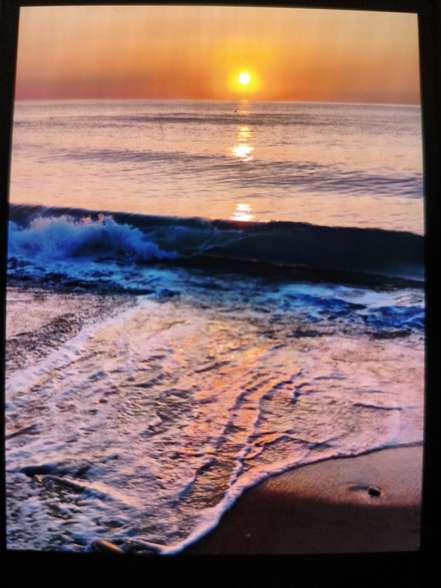 A seascape is depicted under a bright orange sunset. A radiant sun is positioned directly above the horizon, its reflection shimmering across the calm waters of the sea. Below the horizon, a wave is crashing, its form detailed with a mix of blue hues. The shoreline is visible in the foreground with the foamy edge of a wave advancing onto the beach. The wet sand reflects the colors of the sunset.