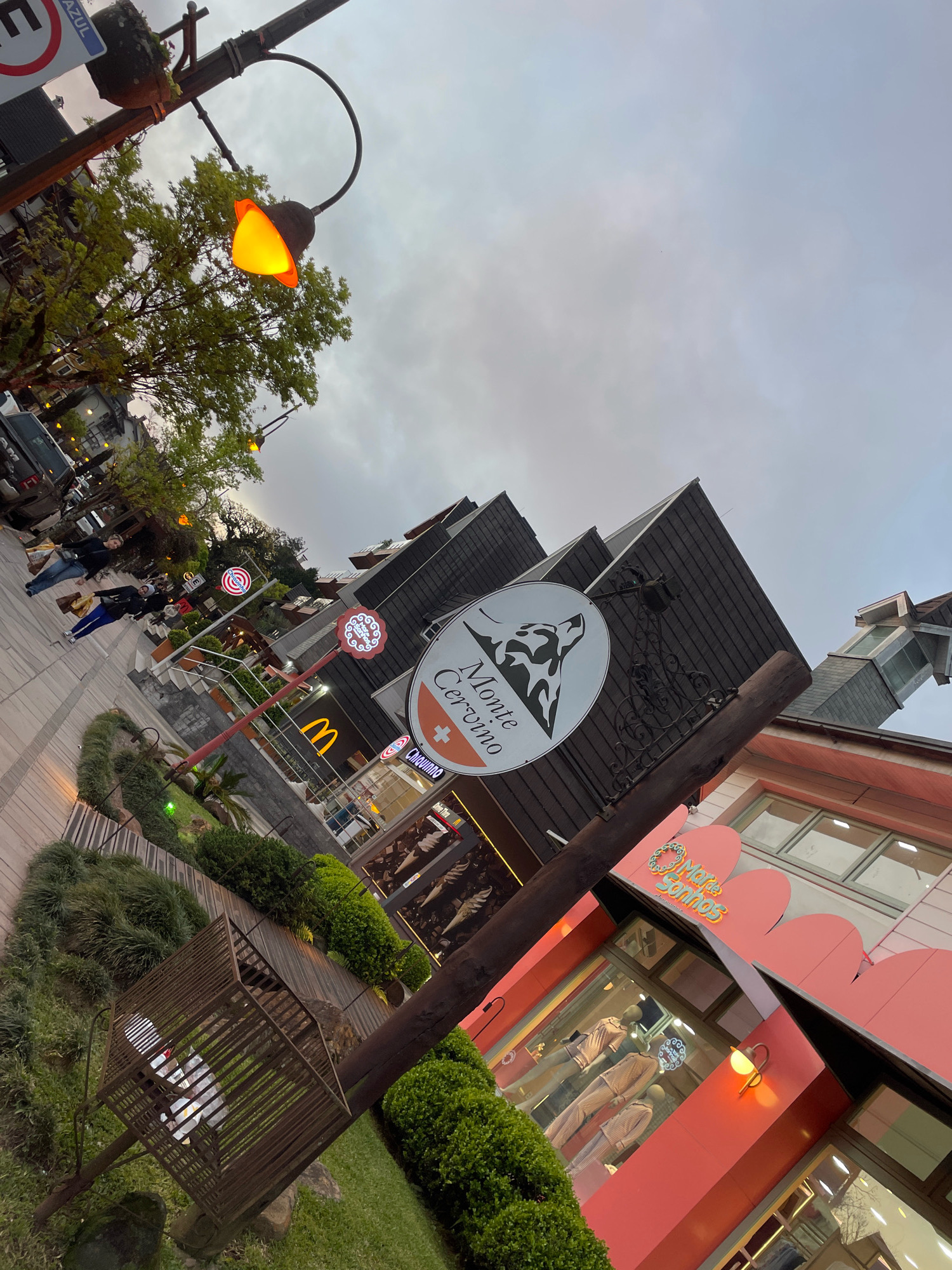 The image captures a street scene in Gramado, Brazil, likely during the evening or overcast day, with a tilted perspective. A prominent lamppost with an orange glowing bulb illuminates the foreground. To the left, a sidewalk features several people walking and carrying shopping bags. Adjacent to the sidewalk, a building with a dark, angled roofline is visible, housing businesses. One sign reads "Monte Cervino" with a wolf logo and a Swiss cross. Another sign, possibly for McDonald's, displays the iconic golden arches. Further to the right, a pink facade building has large windows displaying mannequins dressed in pajamas. A sign above this store reads "Marie Sonhos". Lush green landscaping, including bushes and grass, is present in the foreground and along the sidewalk, with a decorative, cage-like structure for plants. The sky is overcast with a cloudy texture.