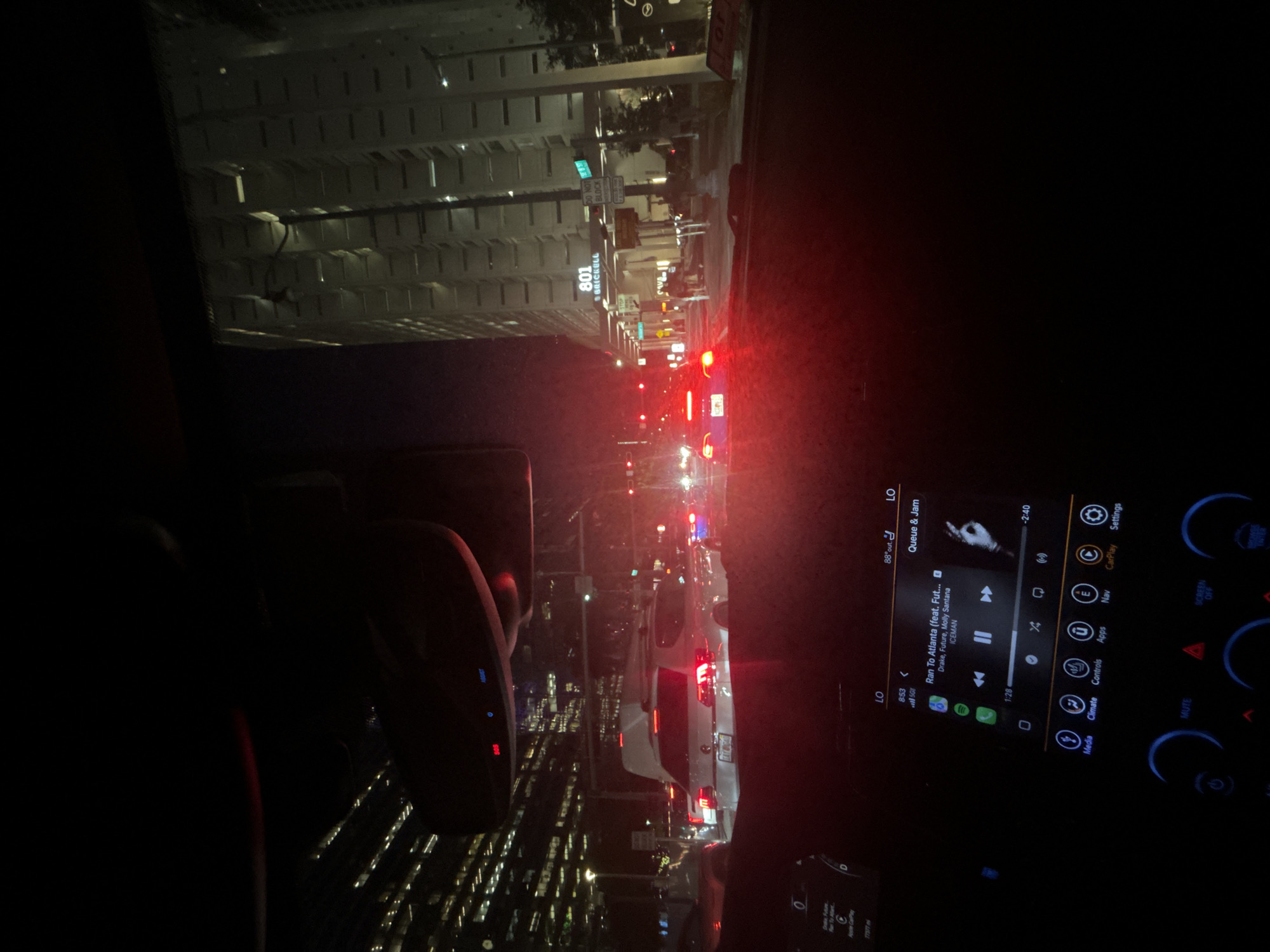 This media file captures a scene from inside a vehicle, likely a car, at night in a city. The view is directed forward, showing traffic and the urban environment. The main subjects are the road, other vehicles, and the illuminated buildings lining the street. A digital display, presumably a car's infotainment system, is visible on the right side of the frame, showing song information and controls for media, climate, and navigation. The song displayed is "Ran To Atlanta" by Drake, Future, and Molly Santana, featuring ICEMAN.
Notable details include the time of day being night, indicated by the darkness and artificial lighting. The weather appears to be clear, as there are no visible signs of rain or fog. Location cues suggest an urban setting, with tall buildings and streetlights. The building on the left is marked with "801 SBRICKELL," indicating a specific location in Miami, United States of America. Traffic lights and brake lights of other cars are prominent, suggesting the vehicle is in traffic. The overall mood is typical of a nighttime drive in a bustling city. The focus is on the sensory experience of being in a car at night, with the interplay of artificial lights and the sounds of music.