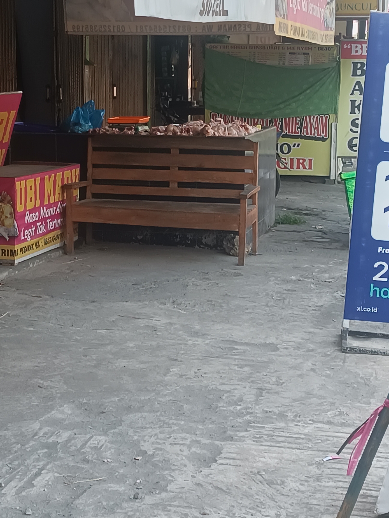 The image shows an outdoor scene in Imogiri, Indonesia, likely a street or market area. In the foreground, a grey concrete ground is visible with some debris. A wooden bench is positioned centrally, with a pile of raw chickens or meat on a wooden counter in front of it. To the left of the bench, a pink box with the text "UBI MADU" is displayed, indicating a food product. In the background, various signs and awnings are visible, some with Indonesian text. A green tarp hangs behind the counter. To the right, a blue sign with white lettering is partially visible, featuring the website address "xl.co.id". The overall impression is of a local food stall or market in a commercial area.