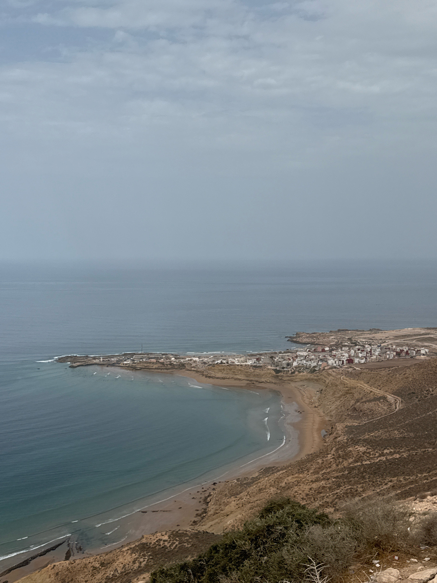 An aerial, eye-level view captures the coastal town of Imsouane, Morocco, with its distinctive bay and the vast Atlantic Ocean under a cloudy sky. The town itself is situated on a rocky peninsula that juts out into the ocean, featuring a dense collection of buildings with white walls and red-tiled roofs. A winding road is visible leading into the town from the right side of the frame, disappearing behind a rocky, arid hillside covered with sparse vegetation. The bay exhibits a crescent shape, with a sandy beach lining its curve. Gentle waves are breaking on the shore, and a few small boats are visible in the water. The ocean's surface is a muted blue-grey, reflecting the overcast sky. The foreground is dominated by the rugged, brown terrain of the hillside, with patches of dry brush and rocks. The overall impression is one of a remote coastal settlement, bathed in soft, diffused light.