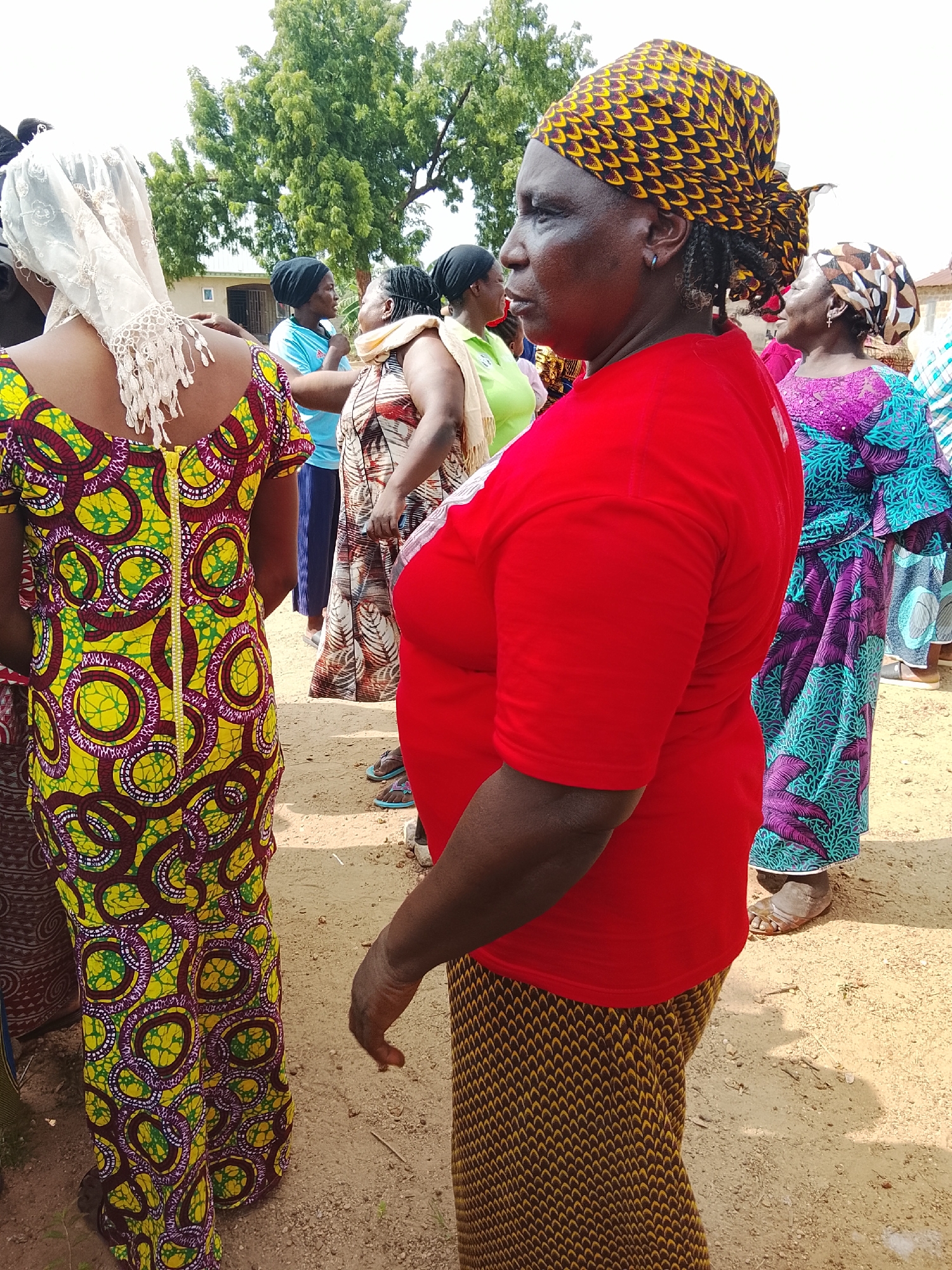 The image captures an outdoor gathering of women in Akwanga, Nigeria. In the foreground, a woman on the right is visible from the side, wearing a vibrant red shirt and a patterned skirt. Her head is adorned with a yellow and black patterned headscarf. To her left, another woman is seen from the back, wearing a colorful yellow, green, and purple patterned dress and a white lace head covering. Other women in the background are also wearing brightly colored African print clothing and headscarves. The ground is sandy and appears to be a dirt path or clearing. Trees with green foliage are visible in the background, suggesting a rural or semi-urban setting. The overall impression is of a community event or social gathering.