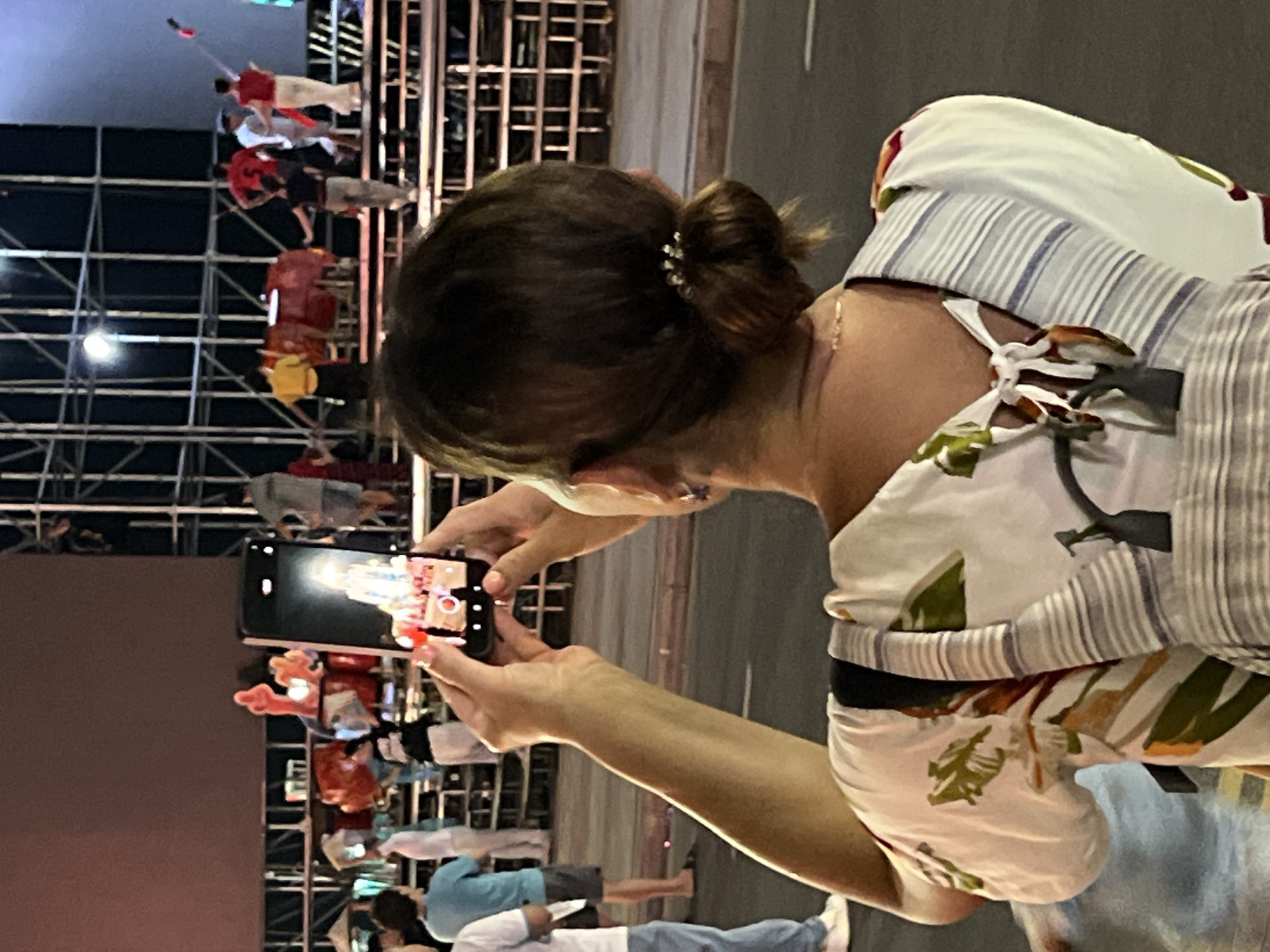 In Nha Trang, Vietnam, a person from behind holds up a smartphone to record a performance taking place on a tiered stage. The stage is adorned with scaffolding and illuminated by bright lights, suggesting a nighttime event. On the stage, several individuals are visible, some in vibrant costumes. Two figures in red shirts and white pants are prominent, one holding a long pole. Other performers are dressed in yellow and red. The background features a large, dark screen, possibly a display for the event. The foreground is occupied by the person filming, whose back is to the camera. They are wearing a patterned top and have their hair tied in a bun. A striped bag is slung over their shoulder. The ground appears to be a paved street, indicating the event might be outdoors in a public space within the city.