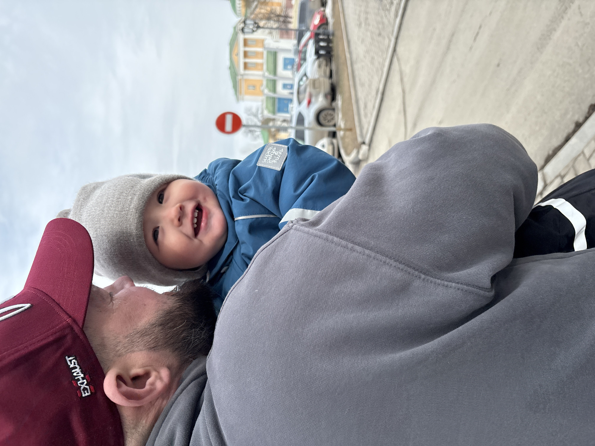 A daytime, eye-level medium shot shows an infant in a blue snowsuit with a gray hat and a man with a red baseball cap lying on the ground in Izhevsk, Russia. The infant, positioned above the man, smiles and appears to be looking at the camera. The man wears a gray hoodie, and the red cap has the word "EXHAUST" embroidered on the side.
In the background, a city street shows cars parked along the curb and a building with colorful facades. A round, red-and-white traffic sign is visible. The sky is overcast. The overall scene suggests a casual, intimate moment between a parent and child in an urban setting.