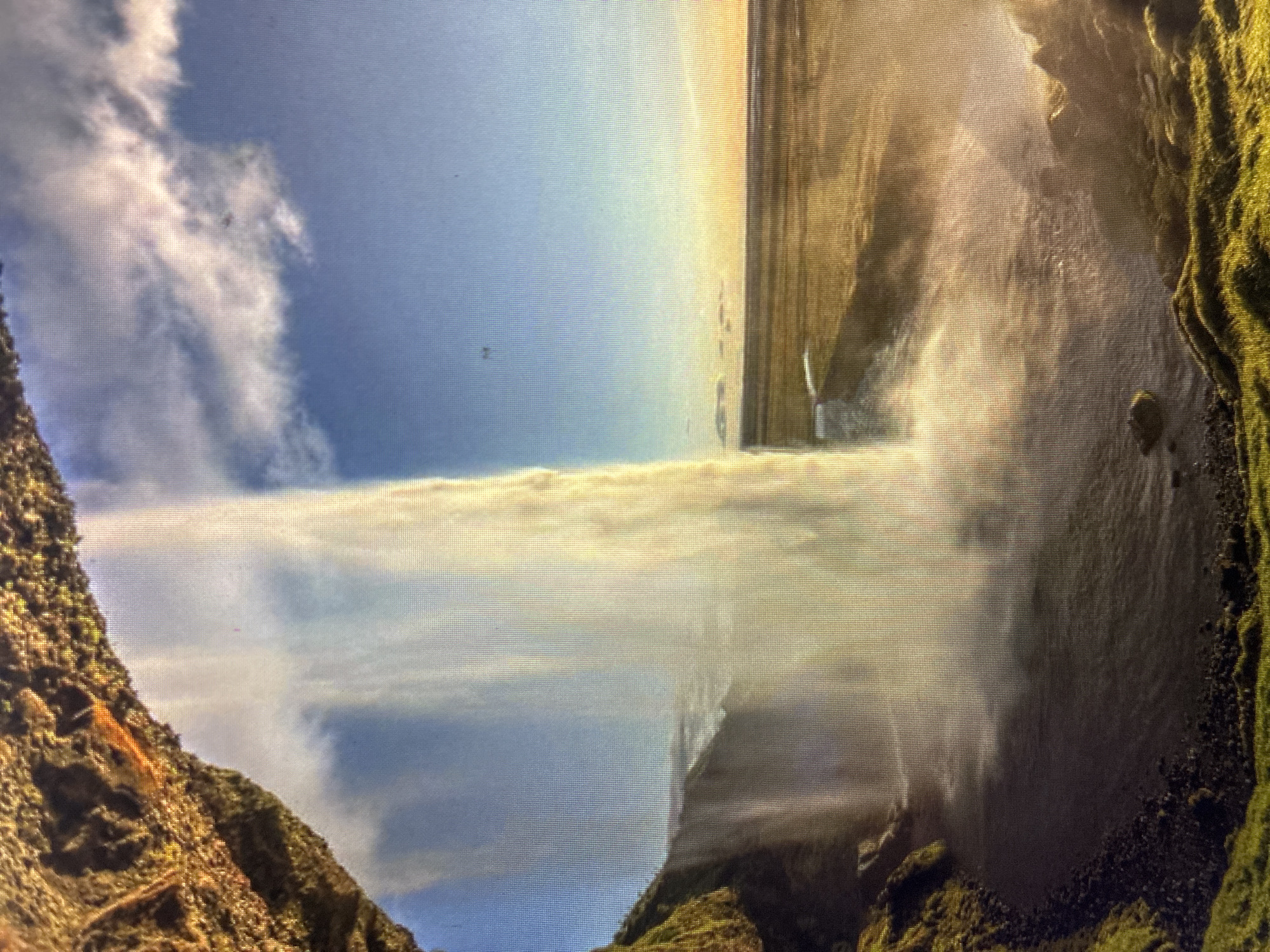 The media file displays a photograph of a majestic waterfall scene, depicting water cascading vertically downwards from a significant height, likely located in the region of Asenovgrad, Bulgaria.
The powerful waterfall dominates the frame, with a large volume of water plunging into a pool or river below. This creates a dense curtain of mist and spray that rises upwards, partially obscuring the lower cliff faces and lending a luminous, ethereal quality to the scene. Steep, rugged cliffs covered in vibrant green moss and vegetation frame the waterfall on both sides, showcasing the natural, untamed environment.
The sky above is bright blue with scattered white, fluffy clouds, indicating clear weather. Bright sunlight illuminates the scene, particularly highlighting the mist of the waterfall and creating various shadows on the rocky terrain. The water at the top of the falls appears to flow from a relatively flat, high plateau or a distinct rock formation before its dramatic descent. The overall impression is one of nature's grandeur and power.
The image itself appears to be a photograph taken of a digital screen, evidenced by a noticeable pixel grid pattern visible across the entire frame. There is no visible text in the image.