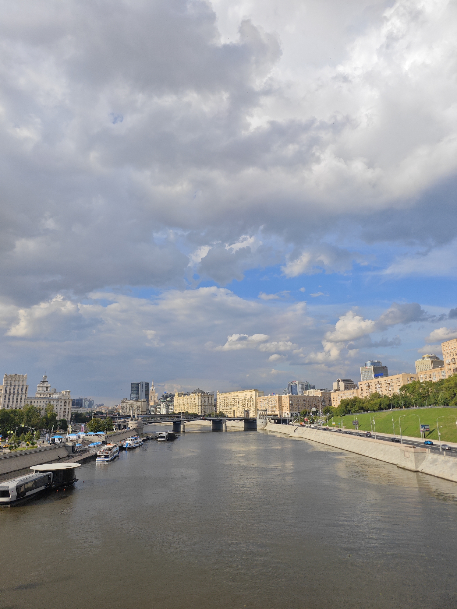 The image captures a wide, dark river flowing through an urban environment. Along the riverbank, a concrete embankment curves into the distance, topped by a road with cars driving. Adjacent to the road is a lush green grassy slope dotted with trees. In the background, a cityscape unfolds, characterized by a collection of buildings of varying architectural styles and heights. These structures include older, ornate buildings with cream-colored facades and newer, more modern high-rises. Several bridges span the river in the distance. Moored along the embankment are several boats, ranging from smaller vessels to larger passenger ferries. The sky above is dramatic, with a mix of dark, heavy clouds and patches of bright blue sky. The overall impression is of a bustling city scene on a potentially overcast day. This scene is consistent with the Moscow River embankment, likely within the central district of Moscow, Russia, given the architecture and the presence of numerous passenger boats characteristic of tourist routes.