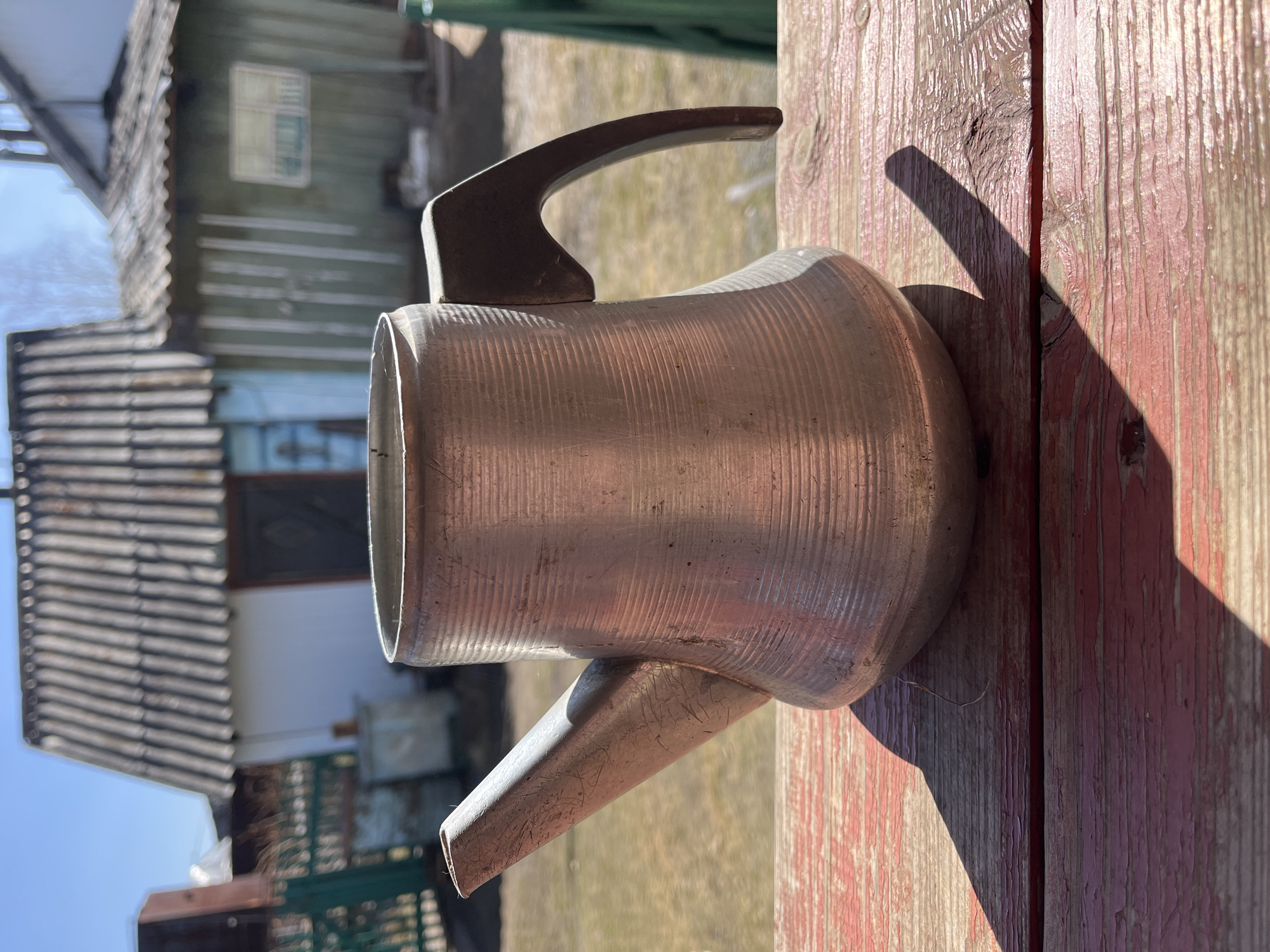 An aluminum watering can is positioned on a weathered wooden surface with red paint in Верхняя Уря, Russia. The can is oriented so that the spout extends downward. The handle is curved and attached to the top of the can. The surface on which the can rests shows distinct linear patterns and texture of wood. In the background, elements of a rural setting are visible, including buildings with shingled roofs and weathered siding. Sunlight casts shadows across the objects and the wooden surface.