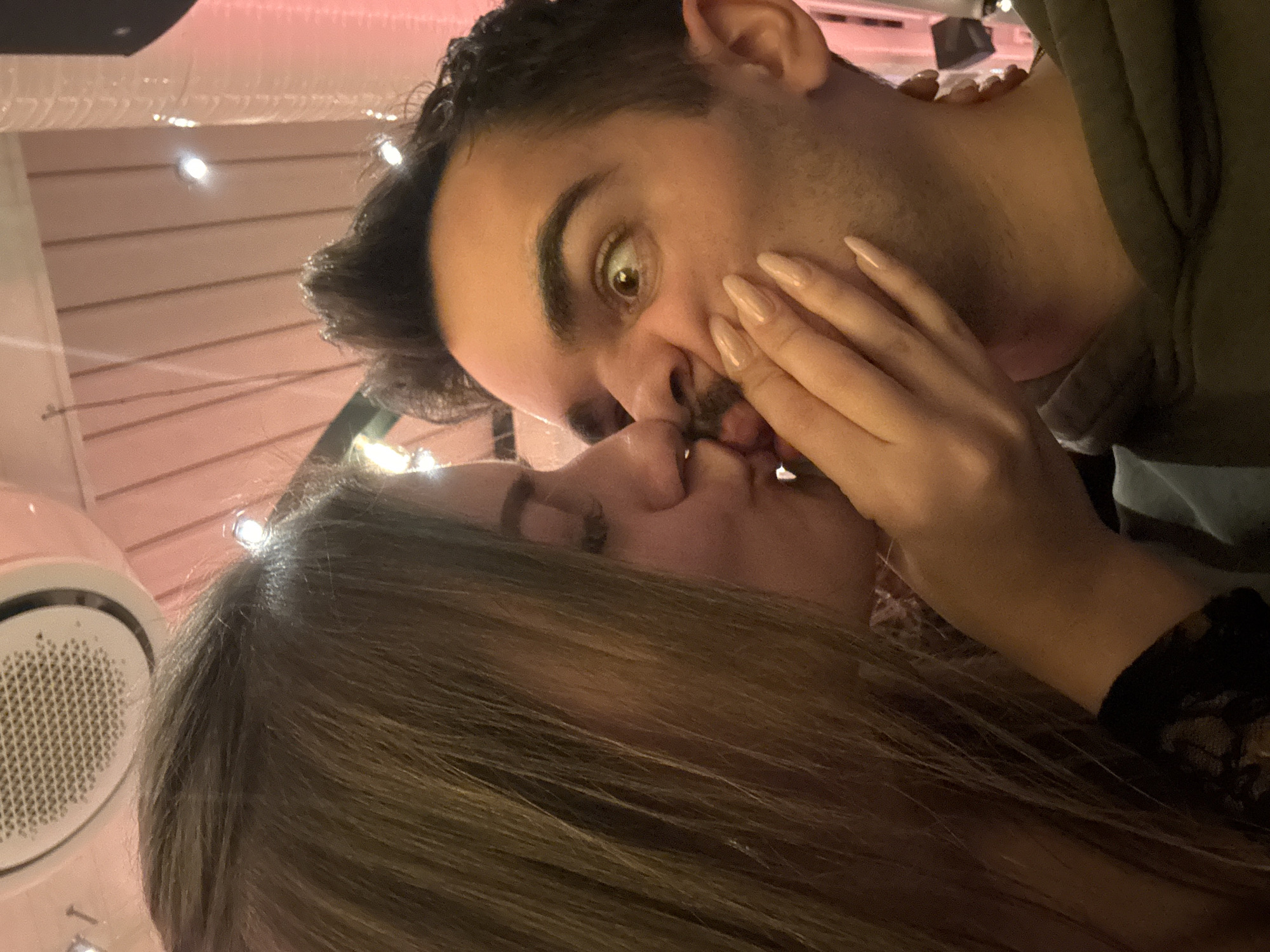 This image depicts an indoor scene in Sofia, Bulgaria, featuring two individuals in close proximity. On the left, a woman with long, brown hair is seen in profile, leaning towards a man. She has closed eyes and pouted lips, appearing to be in the act of kissing the man's mouth. Her right hand, with manicured, light-colored nails, is placed on the man's face, partially pinching his mouth and chin. She is wearing a dark, possibly lace, garment.
On the right, a man is seen from a slightly angled frontal view, looking towards the right side of the frame with a wide-eyed, surprised or humorous expression. His mouth is being pressed by the woman's hand, distorting his facial features. He has dark hair, a light mustache, and stubble. He is wearing a dark olive-green hoodie.
The background is dimly lit with warm, reddish-pink ambient lighting. Visible overhead are ceiling-mounted elements including a circular air conditioning vent with a metallic grid and several small, bright light fixtures. Parallel wooden or paneling slats are also visible on the ceiling and upper wall sections. The overall setting suggests a casual commercial establishment such as a bar or restaurant.