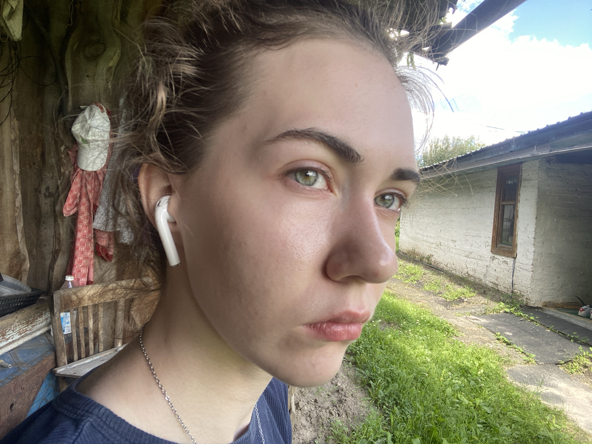 This media file features a close-up portrait of a young woman with light skin and light brown hair. She is wearing a navy blue top and a silver chain necklace. A white wireless earbud is in her right ear. Her expression is neutral, and she is looking to her right.
The background suggests an outdoor, rural or semi-rural setting. To the left, there is a rustic wooden structure with some hanging fabrics and a white cap. To the right, a white brick building with a small wooden window is visible, with a concrete path and overgrown grass leading towards it. The sky is bright with some clouds, indicating it is daytime. The overall atmosphere is casual and appears to be a candid shot.
The location context provided is Zavodoukovsk, Russia. There is no visible text in the image.