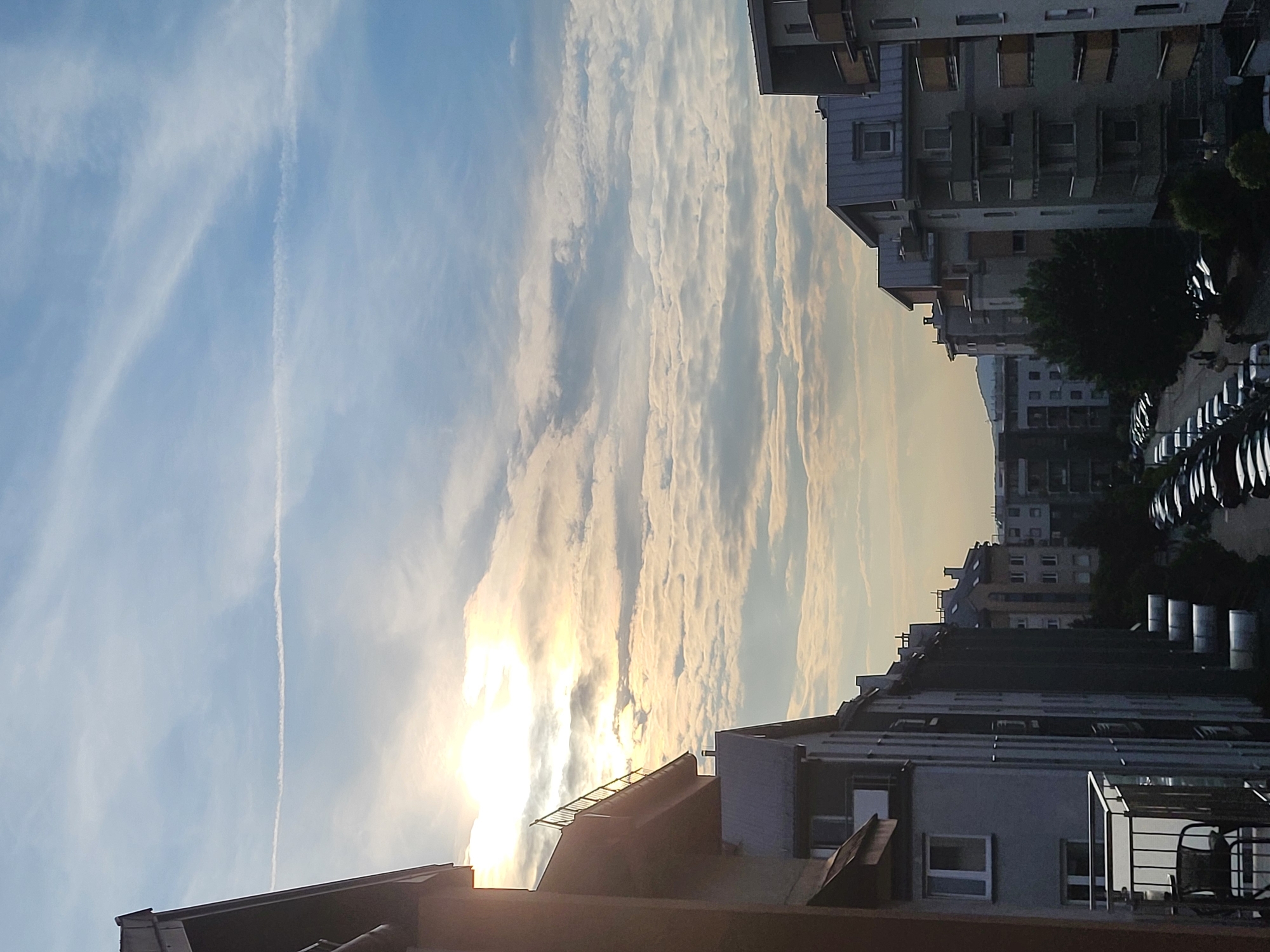 The image depicts a view looking outwards from a balcony or window towards a city street lined with apartment buildings. The sun is visible in the upper left quadrant, partially obscured by clouds, casting a warm glow. A long, thin contrail stretches across the bright blue sky. The foreground shows a part of a building with a balcony railing and outdoor furniture. The middle ground consists of several apartment buildings of varying heights and architectural styles, many with balconies. A street is visible below, filled with parked cars. In the background, more buildings and a distant hill can be seen. The overall scene suggests a residential area within a city.