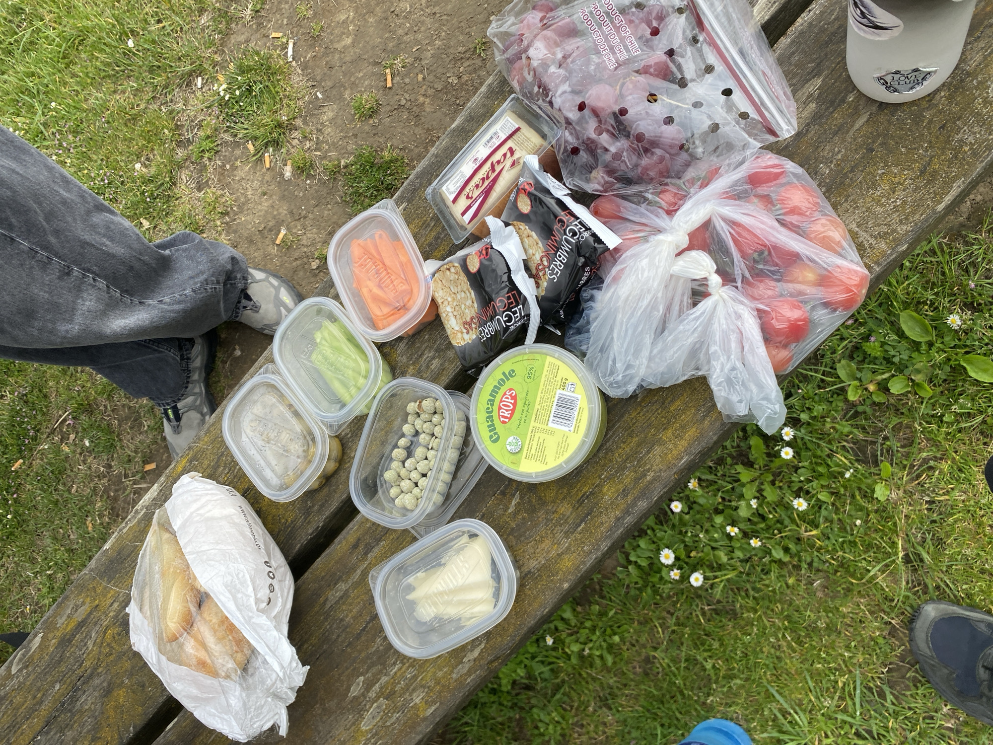 On a weathered wooden bench outdoors in Villaviciosa, Spain, a picnic is laid out. The scene shows food items including:
* Grapes and tomatoes in plastic bags
* A container of guacamole
* Rice cakes
* A block of cheese
* Several clear plastic containers holding carrots, celery, olives, capers, and pieces of cheese.
* A white plastic bag filled with bread.
A water bottle is visible towards the top of the image. The ground nearby is covered in grass, with cigarette butts and small white flowers scattered around. A person's leg wearing jeans and grey sneakers is visible to the left. Another person wearing a shoe can be seen on the bottom right of the image.
