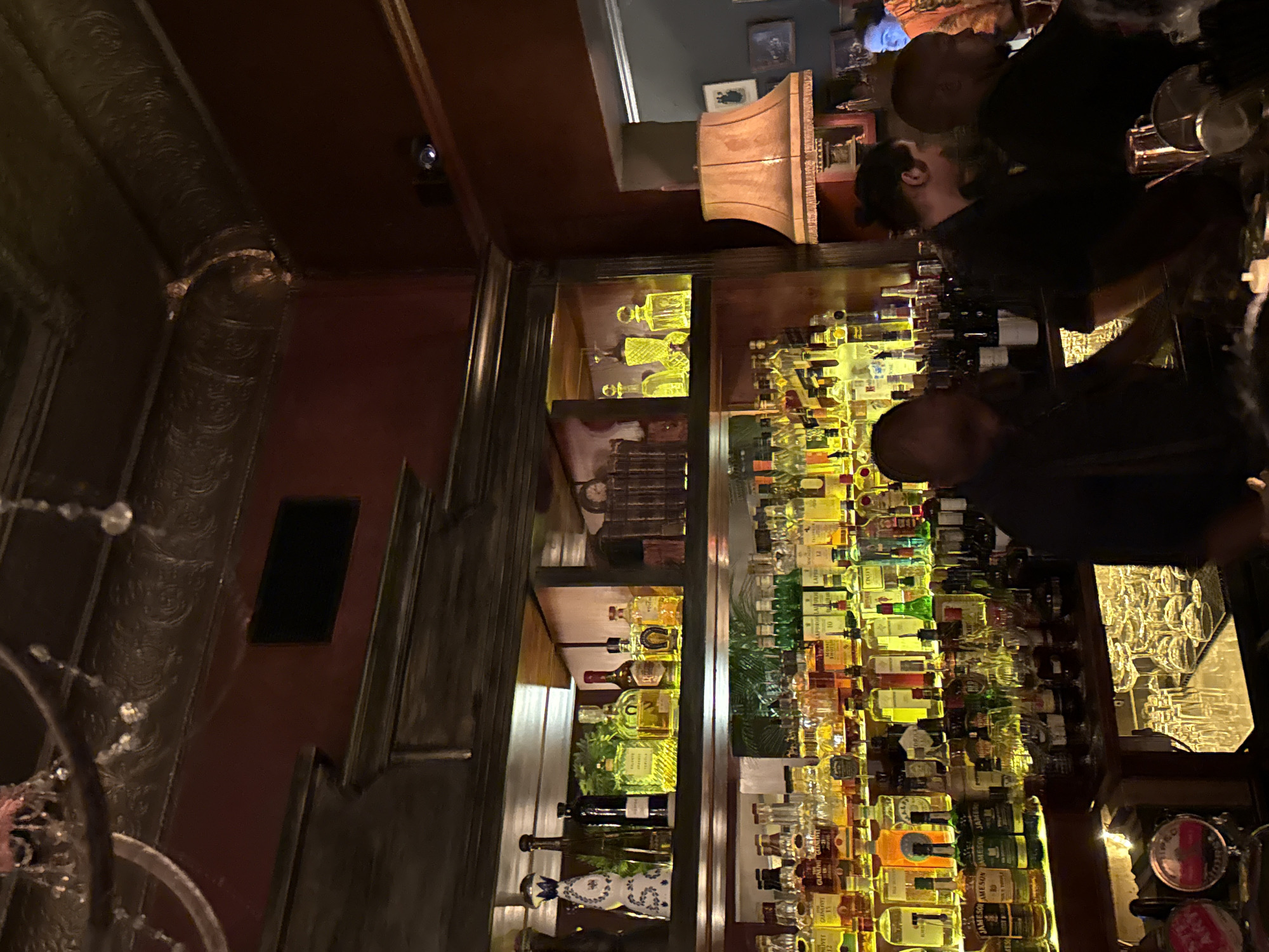This is an interior shot of a bar, possibly in Cape Town, South Africa. The scene shows the back of the bar with a tall, ornate liquor display case filled with bottles of various spirits. There are at least two bartenders in the foreground, engaged in their work, and glasses are visible on the bar surface. A lamp and framed pictures are on the wall behind the bar. The environment is dimly lit, creating a warm ambiance.