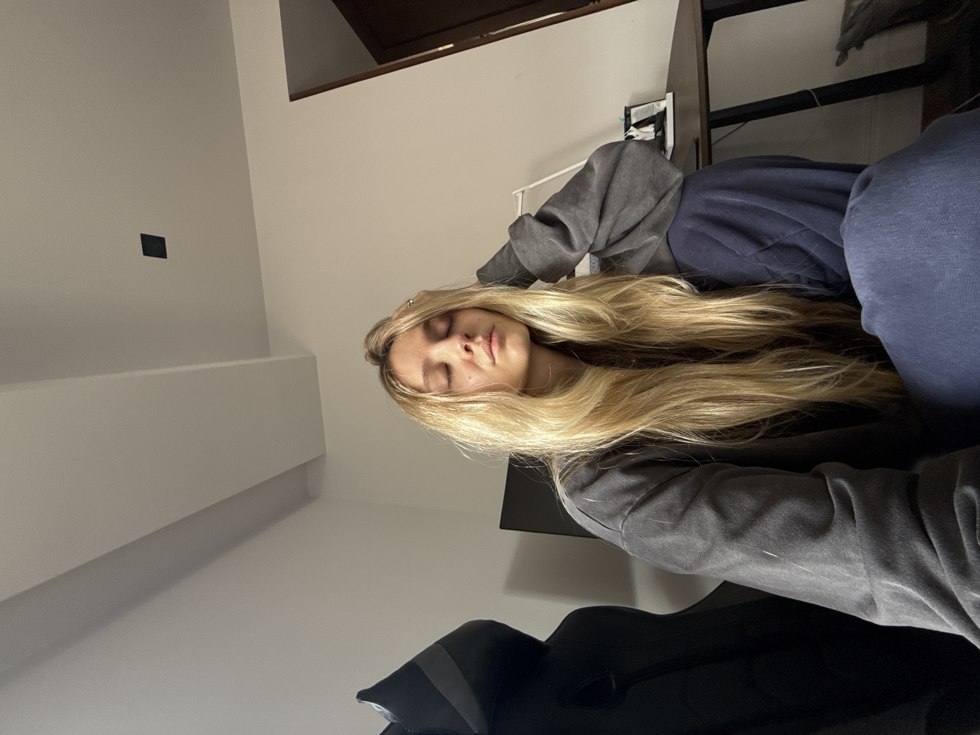 The image displays a young woman with long, wavy blonde hair resting or sleeping indoors. Her eyes are closed, indicating a state of peace, relaxation, or sleep. She is wearing a dark gray hooded sweatshirt and dark blue pants or sweatpants. Her right hand, adorned with a metallic ring, is gently placed on her head amidst her hair.
The setting is an indoor room with light-colored walls. In the upper left portion of the frame, a small, dark square object, possibly a light switch or electrical outlet, is visible on the wall. Above and behind the woman, a dark brown wooden structure, potentially a bed frame, headboard, or part of a shelf, can be seen. Below the woman's head, a dark, flat rectangular object, resembling a screen or panel, is present. A dark, padded piece of furniture, possibly a chair, is partially visible in the bottom right corner of the image. The lighting is soft and ambient, suggesting an indoor environment without strong natural light. There is no visible text in the image. The overall scene conveys a calm and quiet atmosphere.