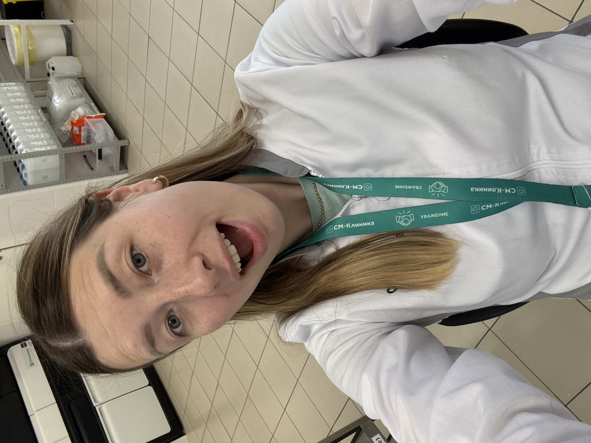 Here's a detailed description of the image:
**General Description:**
The image is a close-up, slightly tilted selfie of a young woman wearing a white medical coat and a green lanyard with "СМ-Клиника" (SM-Clinic) printed on it. She appears to be in a medical setting, possibly a lab or clinic.
**People:**
* **One Woman:** The primary subject is a young woman with light skin, light eyes, and light brown hair. She has an open mouth and a lively expression. She is wearing a gold hoop earring.
**Objects:**
* **Medical Coat:** She is wearing a white medical coat, suggesting she is a healthcare professional.
* **Lanyard:** The green lanyard has the text "СМ-Клиника" (SM-Clinic) and an image of hands shaking.
* **Medical Supplies:** In the background, there is a rack with what appear to be medical supplies or equipment containers, possibly for lab samples.
* **Equipment:** to the left is a medical machine, possibly a blood gas analyzer.
* **Floor:** is lined with square tiles.
**Setting and Location:**
* **Medical Facility:** The background includes elements typical of a medical clinic or laboratory setting. The presence of medical supplies and the woman's attire point to this.
* **City and Country:** Russia, Moscow.
**Actions and Interactions:**
* **Selfie:** The woman is taking a selfie.
* **Expression:** Her open mouth and facial expression suggest she is happy or excited.