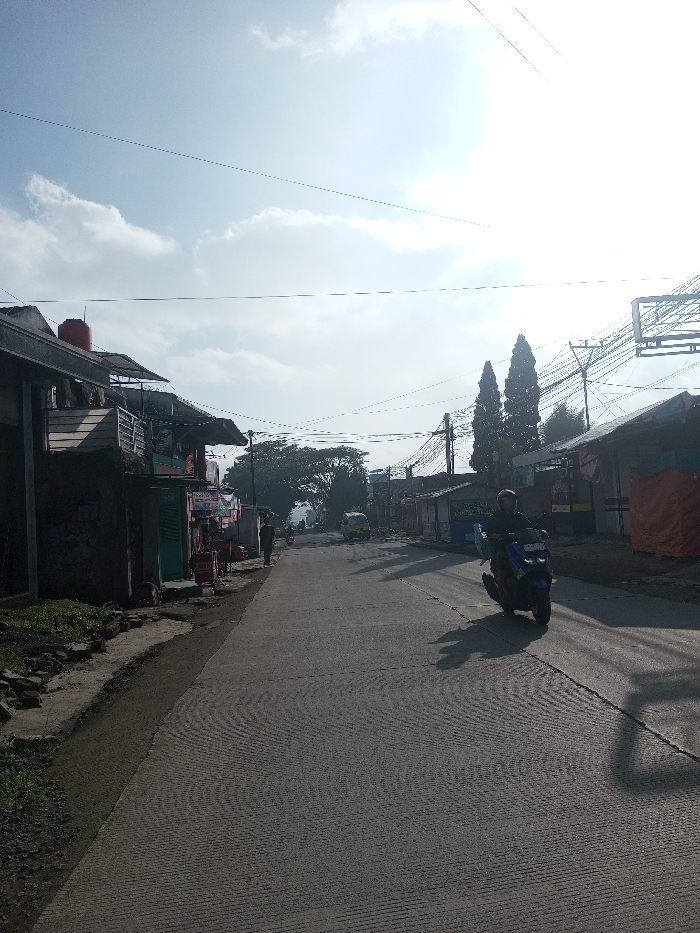 A long, straight road leads into the distance, flanked by modest buildings on either side. Utility poles and wires crisscross overhead. A person rides a scooter toward the viewer on the right side of the road, casting a long shadow. Another person walks along the left side of the street. The sky is bright with scattered clouds. Two tall, slender trees stand in the distance to the right.