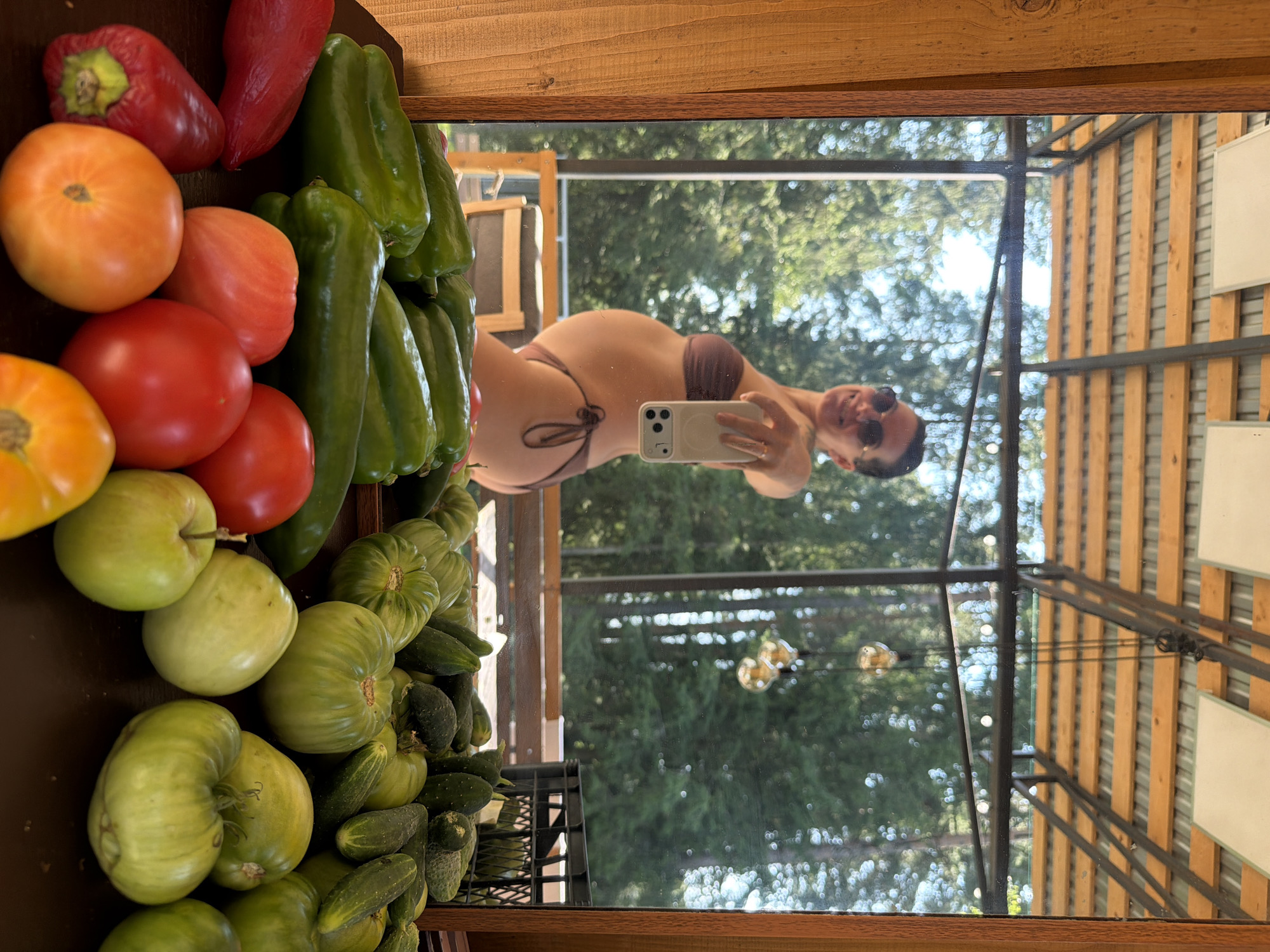 The image shows a pregnant woman in a bikini taking a mirror selfie. She is standing in what appears to be an outdoor covered patio or veranda. In the foreground, on a wooden table, are an assortment of fresh vegetables, including green peppers, red and orange tomatoes, and green tomatoes. Behind the woman, through a large window or glass wall, a forest of green trees is visible under a bright sky. The woman is wearing sunglasses and her hair is pulled back. The reflection in the mirror shows the woman's profile and her pregnant belly. There are light fixtures hanging from the ceiling of the patio.