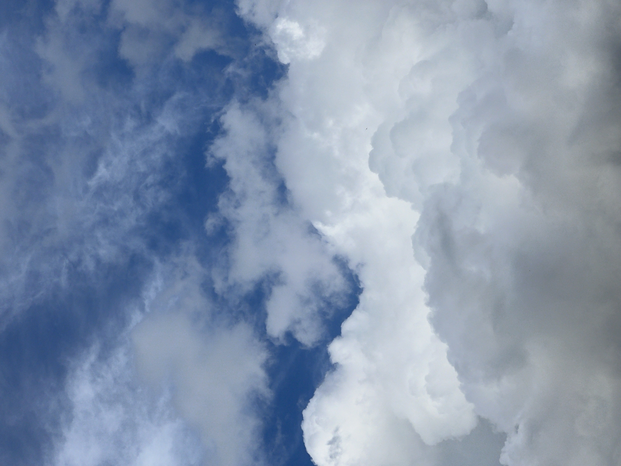 The image shows a view of the sky in an unknown location in an unknown city. The right side of the frame is filled with large, dense, white and gray cumulonimbus clouds. The left side features patches of blue sky with thinner, scattered clouds.