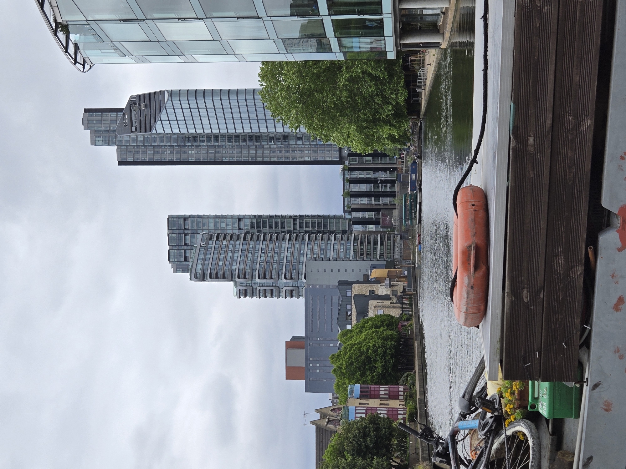 The image depicts a canal scene in London, United Kingdom, with a focus on the waterway and adjacent buildings. The composition is an upward perspective.
To the left of the frame, a line of modern high-rise buildings dominates the view. These structures feature glass facades and varying architectural designs. The buildings are interspersed with patches of green trees and smaller buildings near the canal bank. The sky is overcast.
On the right side of the frame, the edge of a wooden-planked walkway or dock is visible. A bright orange lifebuoy hangs against the side. A bicycle is also partially visible, with its front wheel and handlebars prominent near the lower right corner. The canal water reflects the buildings and sky.