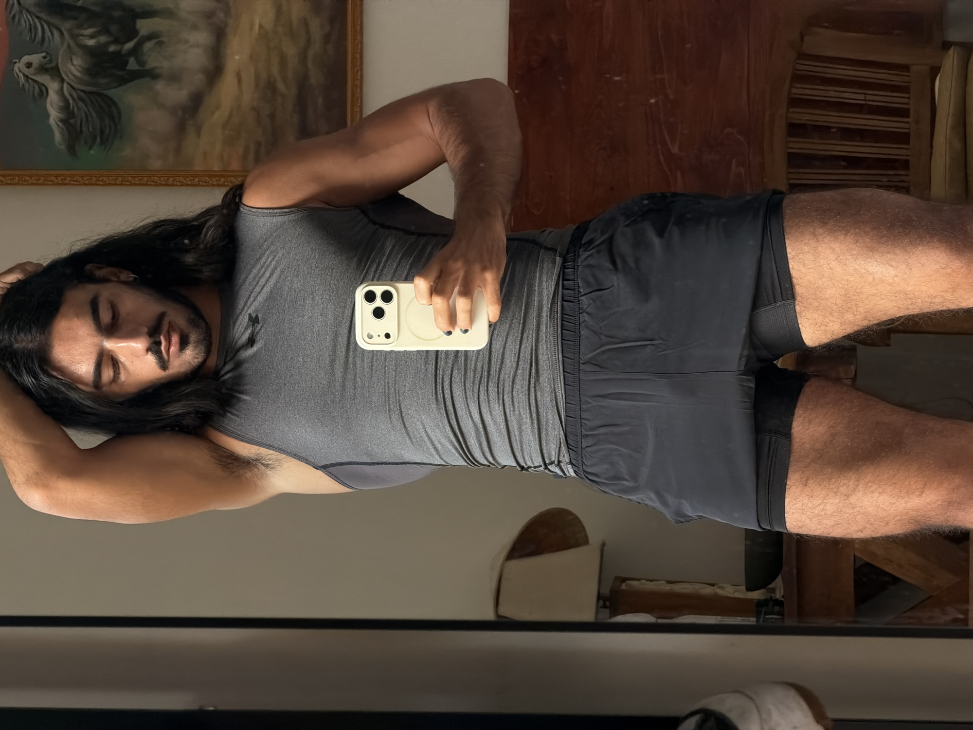 A person with long dark hair and a mustache is taking a mirror selfie in a room. They are wearing a gray tank top and black athletic shorts. The person is holding a white smartphone in their right hand. In the background, there is a lamp, a painting of horses, and a wooden chair. The room appears to be a bedroom. The location is described as being in the city of North Kuta, Indonesia.