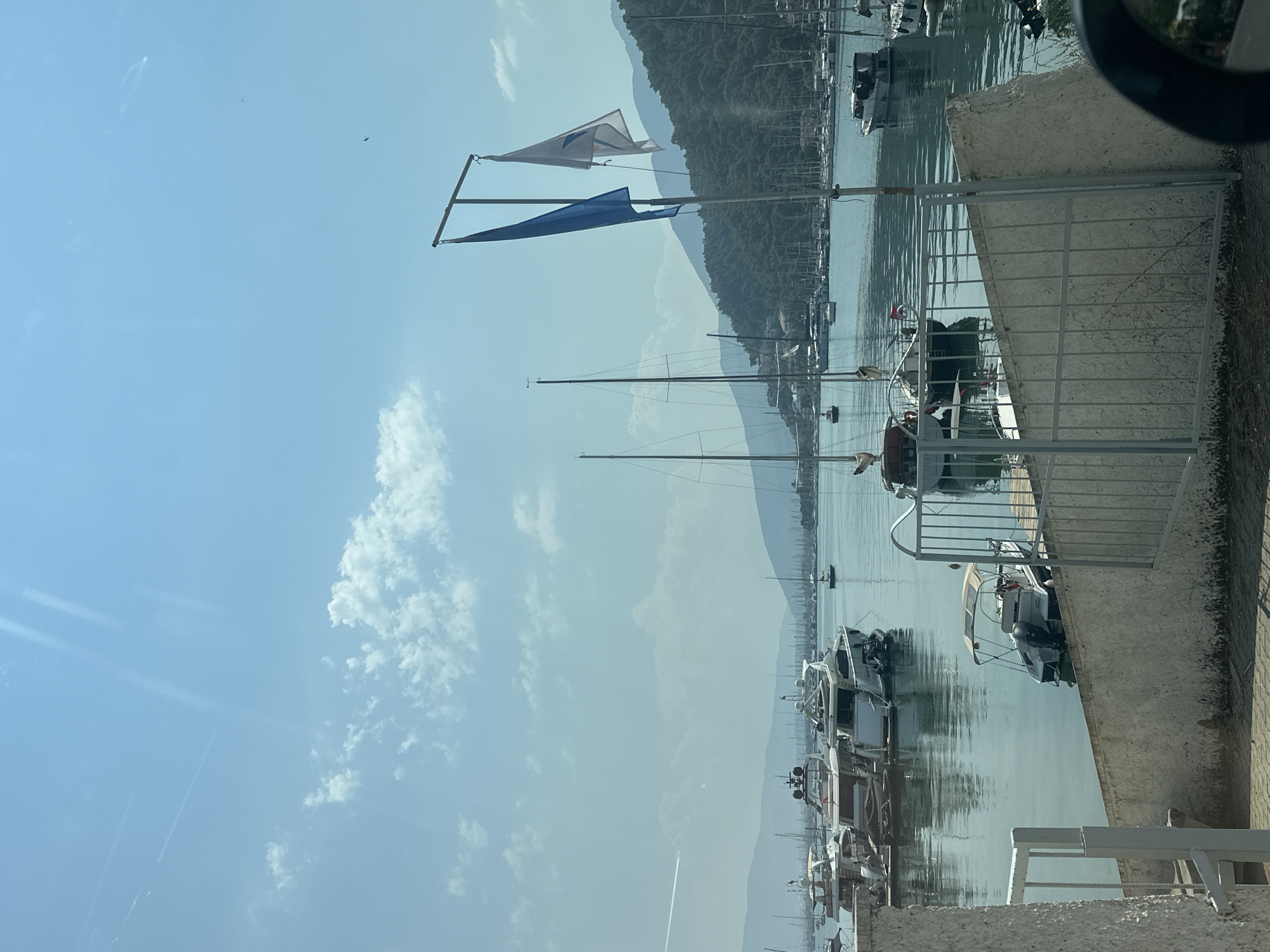 The image depicts a harbor scene in Fethiye, Turkey, bathed in bright daylight under a clear blue sky with scattered clouds. The foreground features a textured white wall and a metal gate, with a cobblestone-like surface visible beneath.
Dominating the middle ground is a serene body of water filled with a variety of vessels. Several large, luxurious yachts are docked, showcasing white hulls and sleek designs. In the distance, a multitude of sailboats and smaller boats are anchored, their masts creating a delicate silhouette against the backdrop of lush, green hills that form the town's natural boundary. A small, open-top motorboat is also visible, adding to the diverse maritime activity.
Two flags, one bright blue and the other white with a crest, are prominently displayed on a flagpole in the mid-right of the frame. The overall atmosphere is one of tranquility and a thriving port. The background is characterized by the imposing, verdant mountains that characterize the landscape of Fethiye. The water reflects the sky and surrounding boats, adding a sense of depth and calm to the scene.