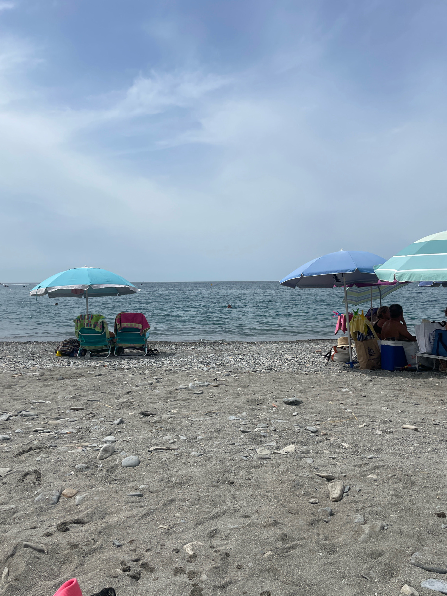 The image depicts a pebble and sand beach in La Herradura, Spain, with the Mediterranean Sea in the background under a cloudy sky. On the left, two empty beach chairs are set up under a light blue umbrella, each draped with a towel. To the right, a group of people are seated under blue and striped umbrellas, with coolers and bags nearby. The sea appears calm with a few swimmers visible in the distance. The overall scene captures a relaxed day at the beach.