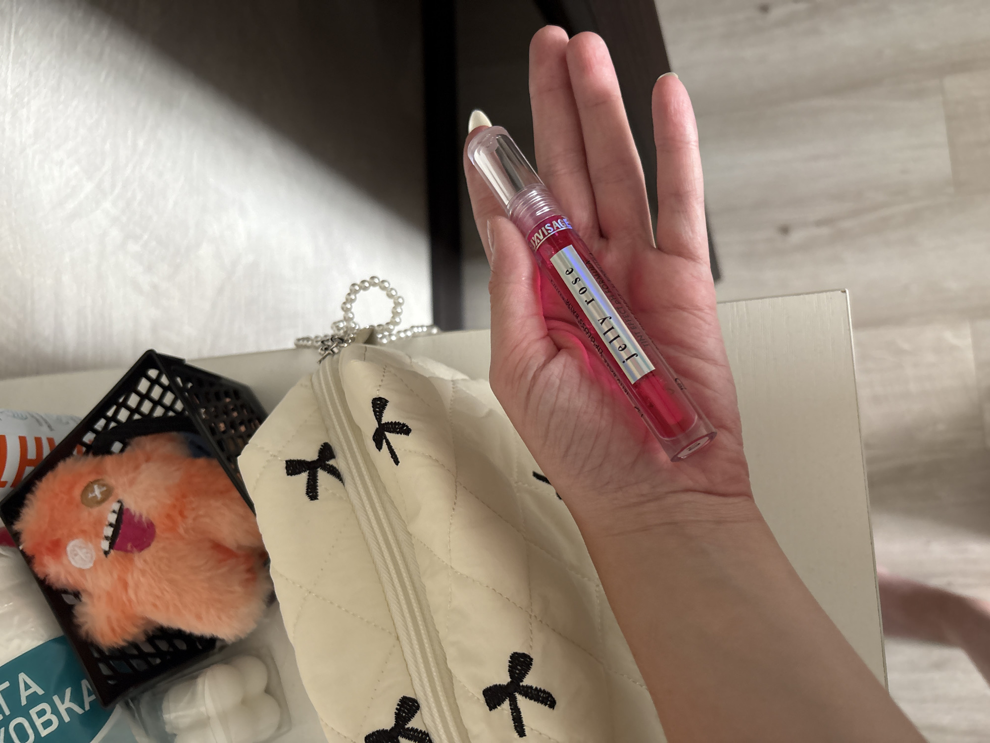 A person's hand is holding a clear tube of pink lip gloss with the label "Jelly Rose" and "Tint Effect pH Formula." The lip gloss is positioned over a quilted cosmetic bag with black bow embroidery. In the background, a fuzzy orange toy with a tongue sticking out sits in a black basket. The scene appears to be indoors, possibly on a dresser or vanity. The context of Minsk, Belarus is not discernible from the image content.