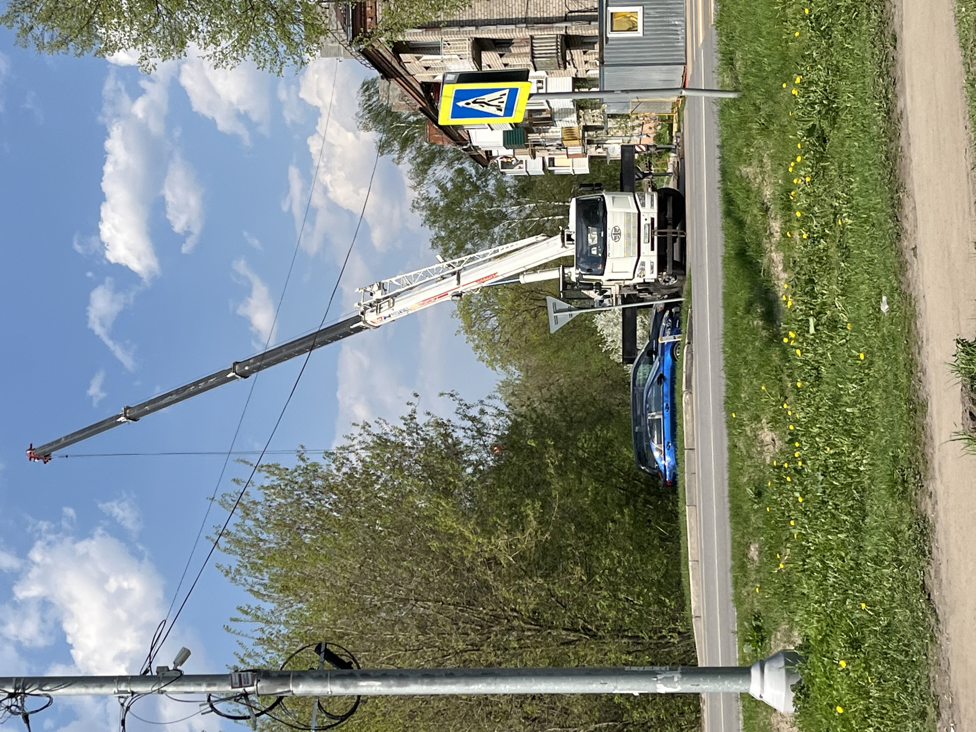 A white crane truck with an extended arm stands on the side of a paved road in Noginsk, Russia. A pedestrian crossing sign is visible behind the crane. A blue car is parked near the crane. A grassy area with yellow flowers lines the road. In the background, there is a building and trees. A pole with wires and a camera is visible in the foreground. The sky is blue with white clouds.