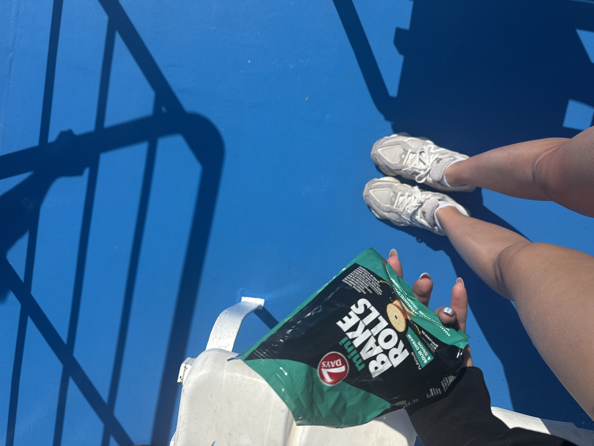The image shows a person's legs and feet wearing white athletic shoes on a bright blue surface, possibly the deck of a boat or a painted outdoor area. The person is holding a green and black bag of "7 DAYS mini BAKE ROLLS" with a "Sour Cream & Onion" flavor. Shadows from what appear to be railings or an overhead structure are cast across the blue surface, suggesting an outdoor setting in sunlight. The background is a vibrant blue, with some white objects visible to the left. The overall impression is a casual outdoor scene, potentially in a sunny location like a coastal area in Greece, given the mention of Prinos.
