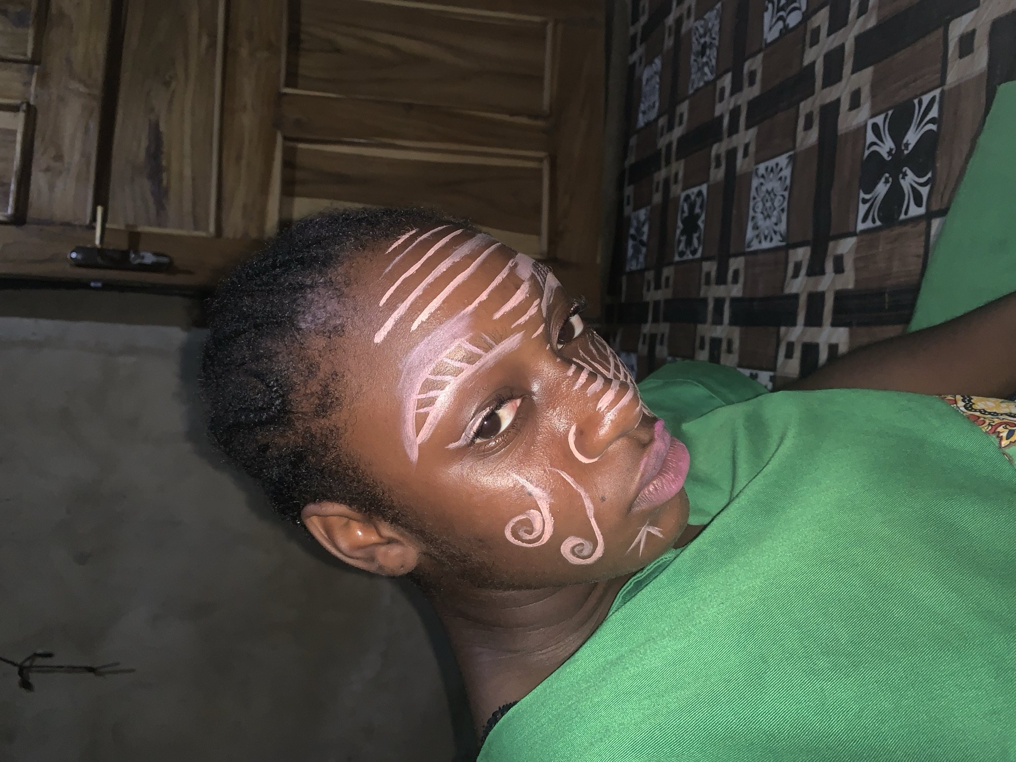A medium shot captures a young Black person adorned with white face paint, looking downwards. The face paint features parallel lines across the forehead and temples, swirling patterns on the cheeks, and a small V-shape below the lip. The individual wears a bright green garment and is positioned against a dark, textured wall. To the right, a wooden door with horizontal panels is visible. The foreground shows a patterned surface with brown, black, and white squares. The overall lighting is dim, suggesting an indoor setting. The location is identified as Agban, Nigeria.