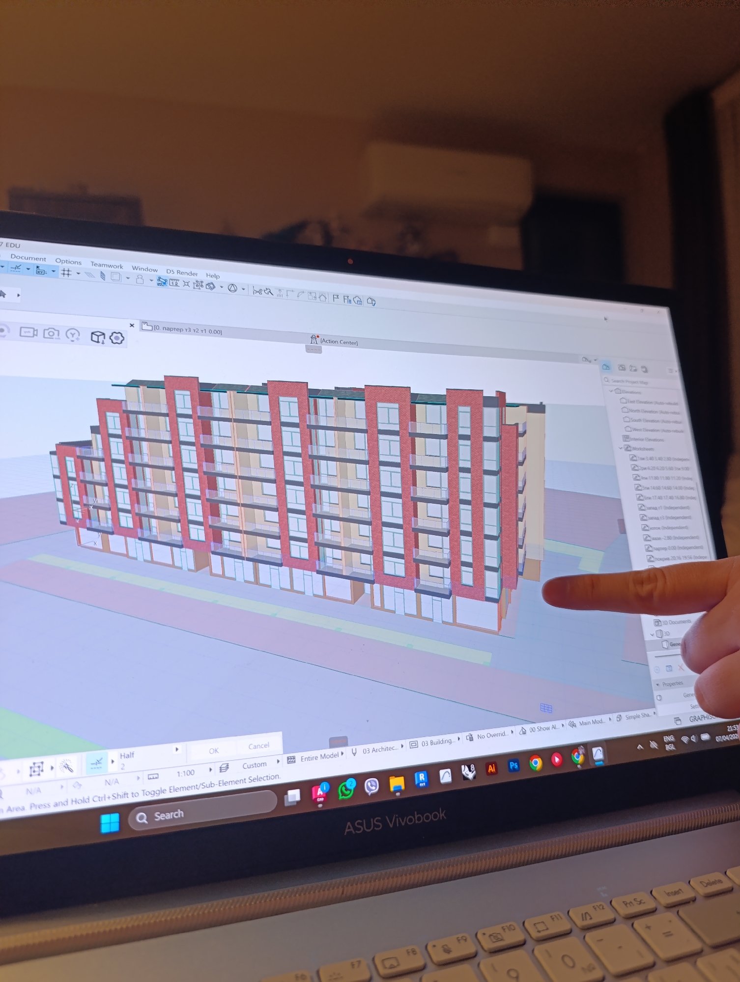 The image shows a laptop screen displaying a 3D architectural rendering. A finger points towards the model, which appears to be a multi-story residential building with a red and white facade, balconies, and numerous windows. The laptop is an ASUS Vivobook. The screen also shows the Archicad software interface. Based on the taskbar, the OS is Windows. There are icons of different programs, including Adobe products. The language setting shows "ENG BGL", which is English and Bulgarian. The time is 21:53 on 07/04/2024. Due to the software's language being set to Bulgarian, it can be inferred that the location may be in Sofia, Bulgaria. The architectural model on screen may represent the design of an apartment building or mixed-use development in Sofia.