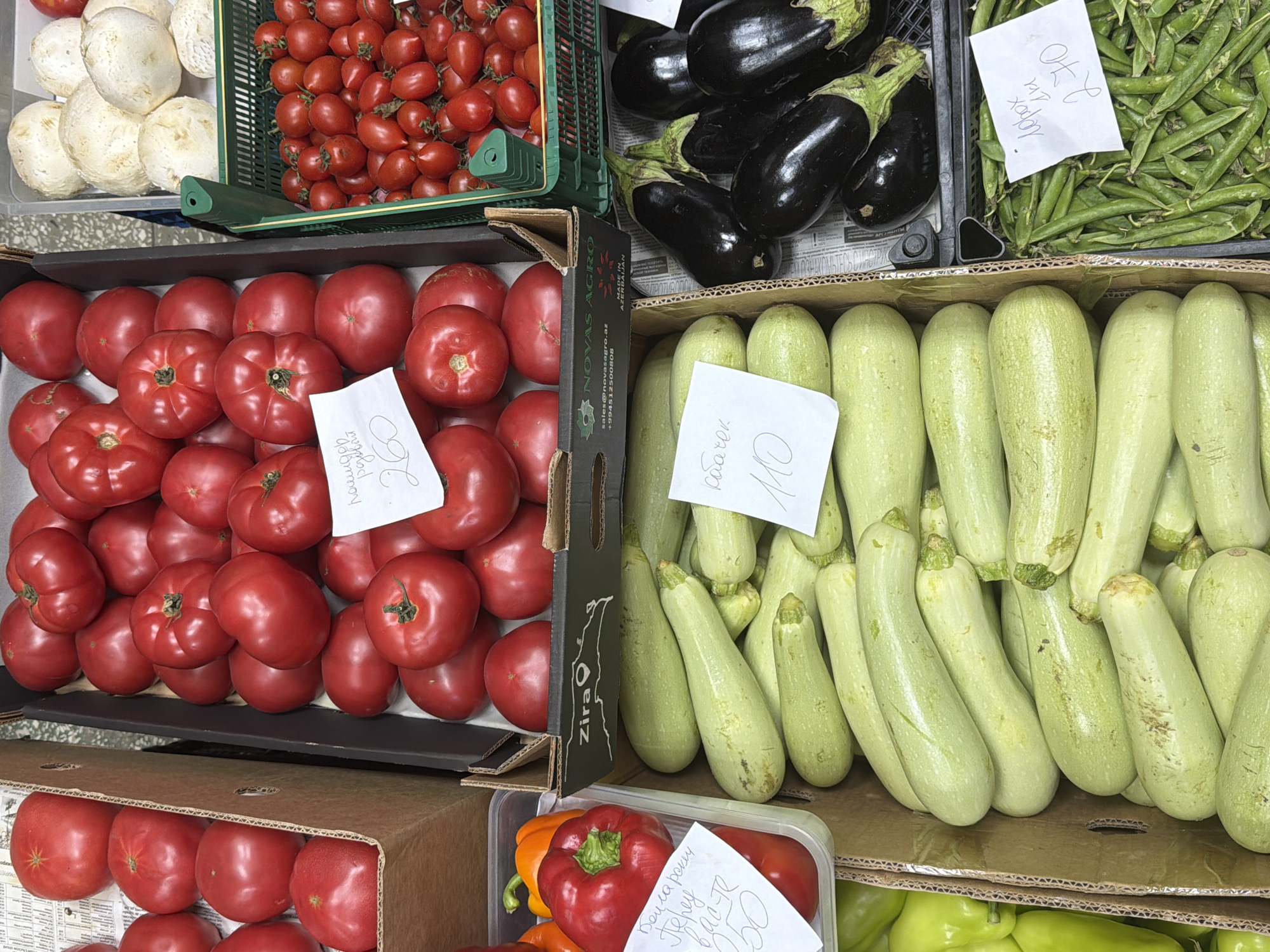 This image shows a street market stall in Sochi, Russia, displaying a variety of fresh produce. In the foreground, large, light-green zucchini are piled high in a cardboard box. To the right of the zucchini, a black plastic crate is filled with dark purple eggplants. Further to the right, a green plastic crate overflows with green peas in their pods.
Behind the zucchini, a cardboard box contains red tomatoes. Another box, to the left of the tomatoes, holds red and orange bell peppers. In the background, a green plastic crate is filled with small red cherry tomatoes, and a gray container holds large white mushrooms. Several price tags are visible, with handwritten prices in what appears to be Russian currency. The overall scene depicts a vibrant market setting with an abundant display of fresh fruits and vegetables.