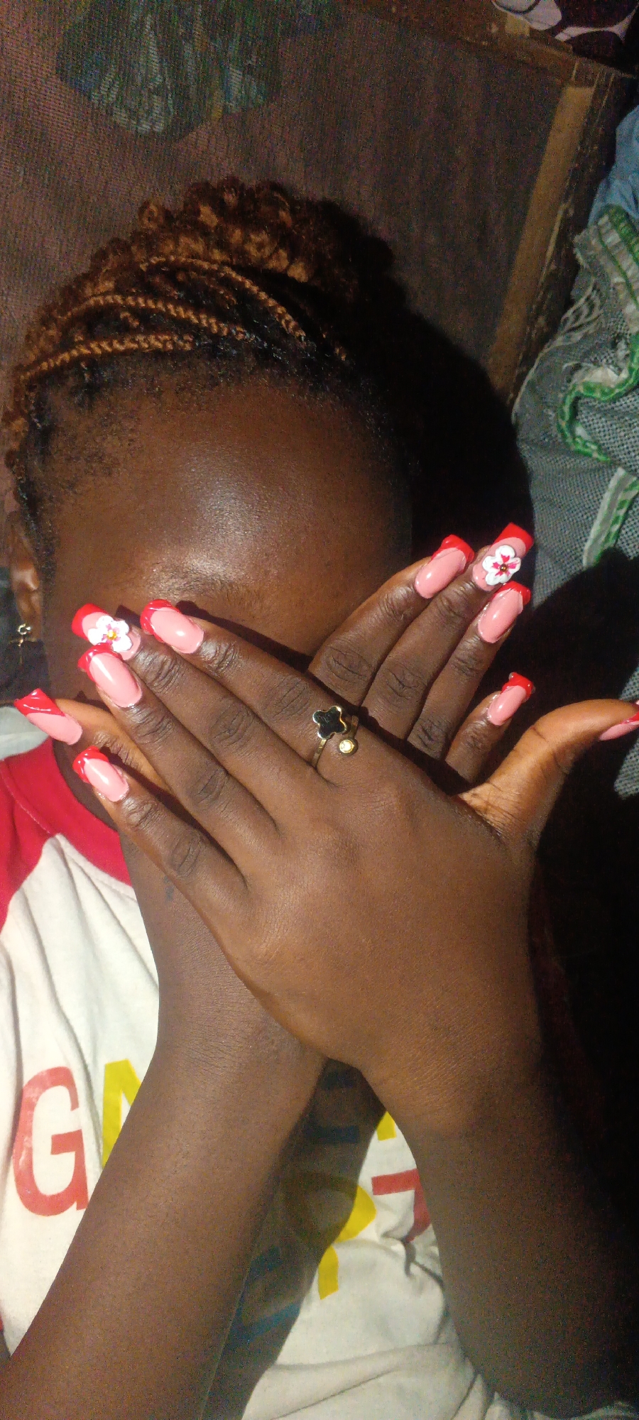 A medium shot captures a person with dark skin and brown braided hair. The person's face is mostly obscured by their hands. The fingernails are long with pink bases and red tips; one nail has a small, white floral design. A gold ring with a black clover-like design and a small pearl adorns one finger. The person is wearing a t-shirt with multicolored lettering. The background is dark and indistinct, showing a bed covered with a green and white striped blanket. The picture was taken indoors. The location of the image is unknown; however, it is mentioned that the location is Onitsha, Nigeria.