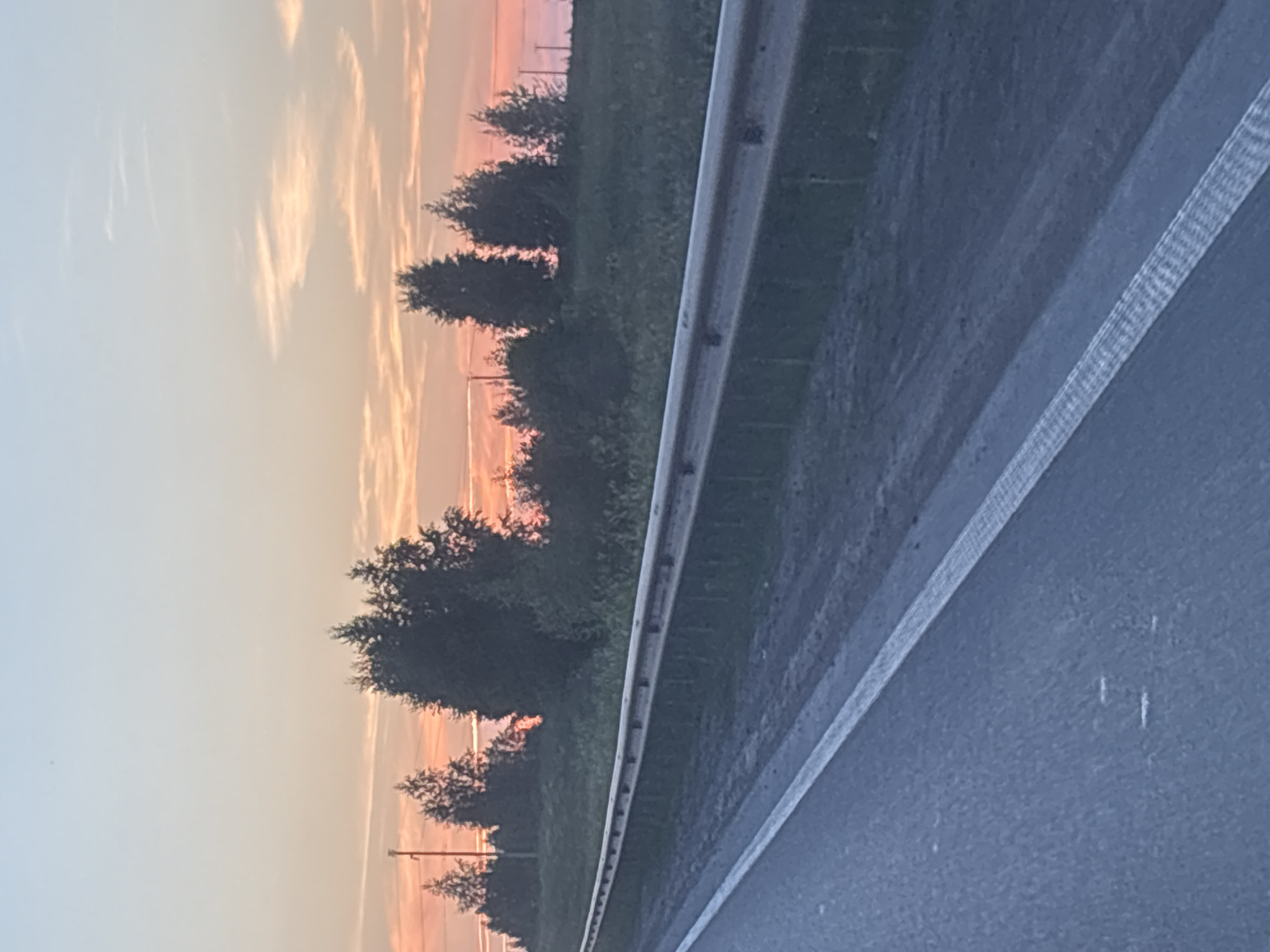 The image depicts a highway scene at sunset, likely in the vicinity of Kazan, Russia, given the foliage and sky coloration often seen in the region during this time. The foreground is dominated by an asphalt road with a white lane divider. To the right of the road, a metal guardrail runs parallel to the pavement, separating it from a grassy embankment. Beyond the embankment, a line of trees and bushes are silhouetted against a sky transitioning from pale blue at the top to hues of orange and pink near the horizon, indicating the sun has recently set or is about to set. The sky is streaked with thin, wispy clouds. Power lines and poles are visible in the distance, peeking through the trees on the left side of the image. No people or vehicles are present. The scene is serene and quiet, characteristic of a rural or semi-rural highway at dusk.