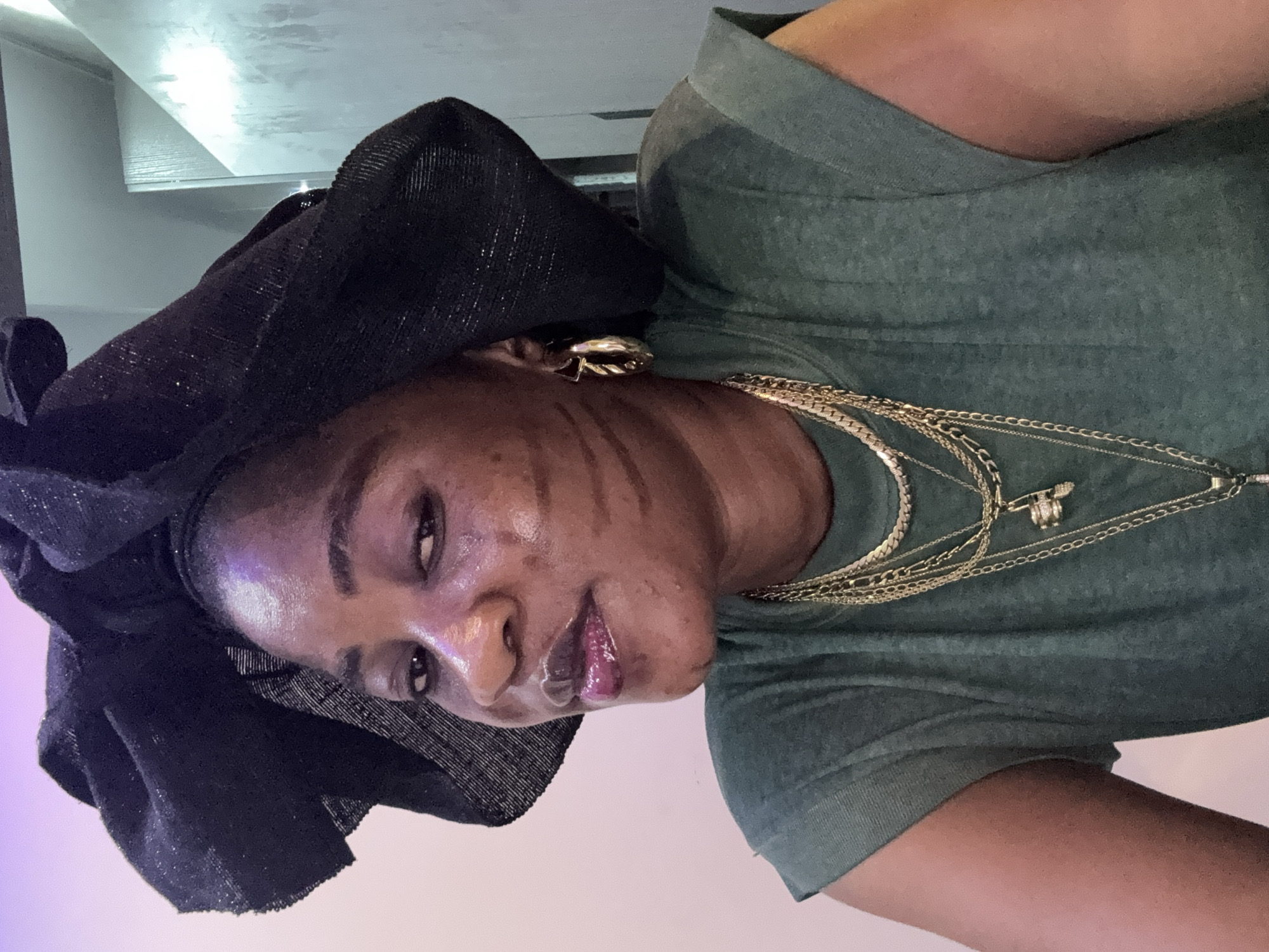 The media file depicts a close-up, head-and-shoulders shot of a woman in Ajara, Nigeria. She is wearing a black headwrap and a green t-shirt. Multiple gold necklaces are layered around her neck. She has dark skin and appears to be wearing makeup, including lip gloss and what looks like body paint or lines drawn on her cheeks. Her expression is directed towards the camera, with her eyes open and lips slightly parted. The lighting suggests an indoor setting, possibly artificial light, with a hint of purple light on the left side of the frame. The background is blurred, with indistinct shapes and textures, including what appears to be a reflective surface above and to the left of the subject. There is no visible text in the image. The overall mood conveyed is one of self-presentation or a casual portrait.